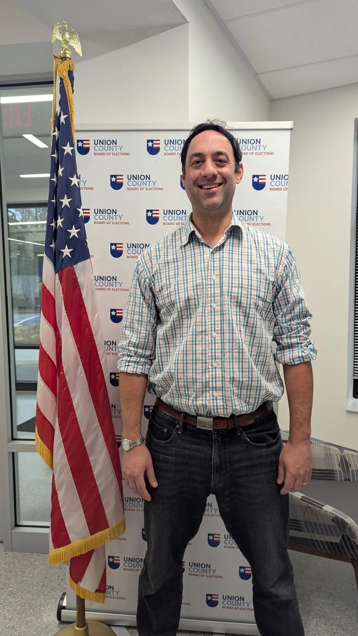 A man standing next to an American flag with a Union County Board of Elections backdrop in the background, smiling at the camera.