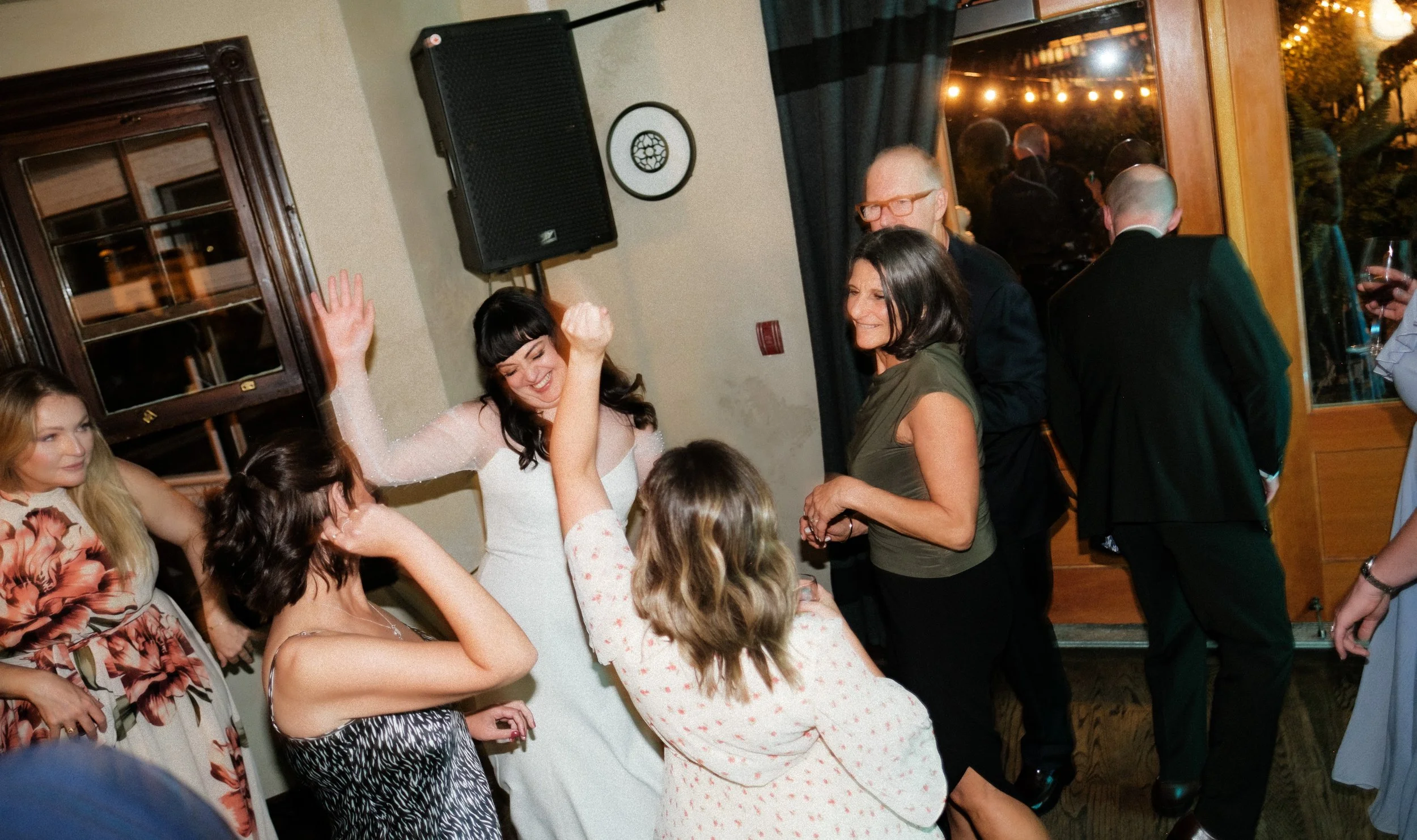 People dancing and enjoying at a party indoors, with some holding drinks, near a doorway with a glass window and curtains, in a warm-lit room.