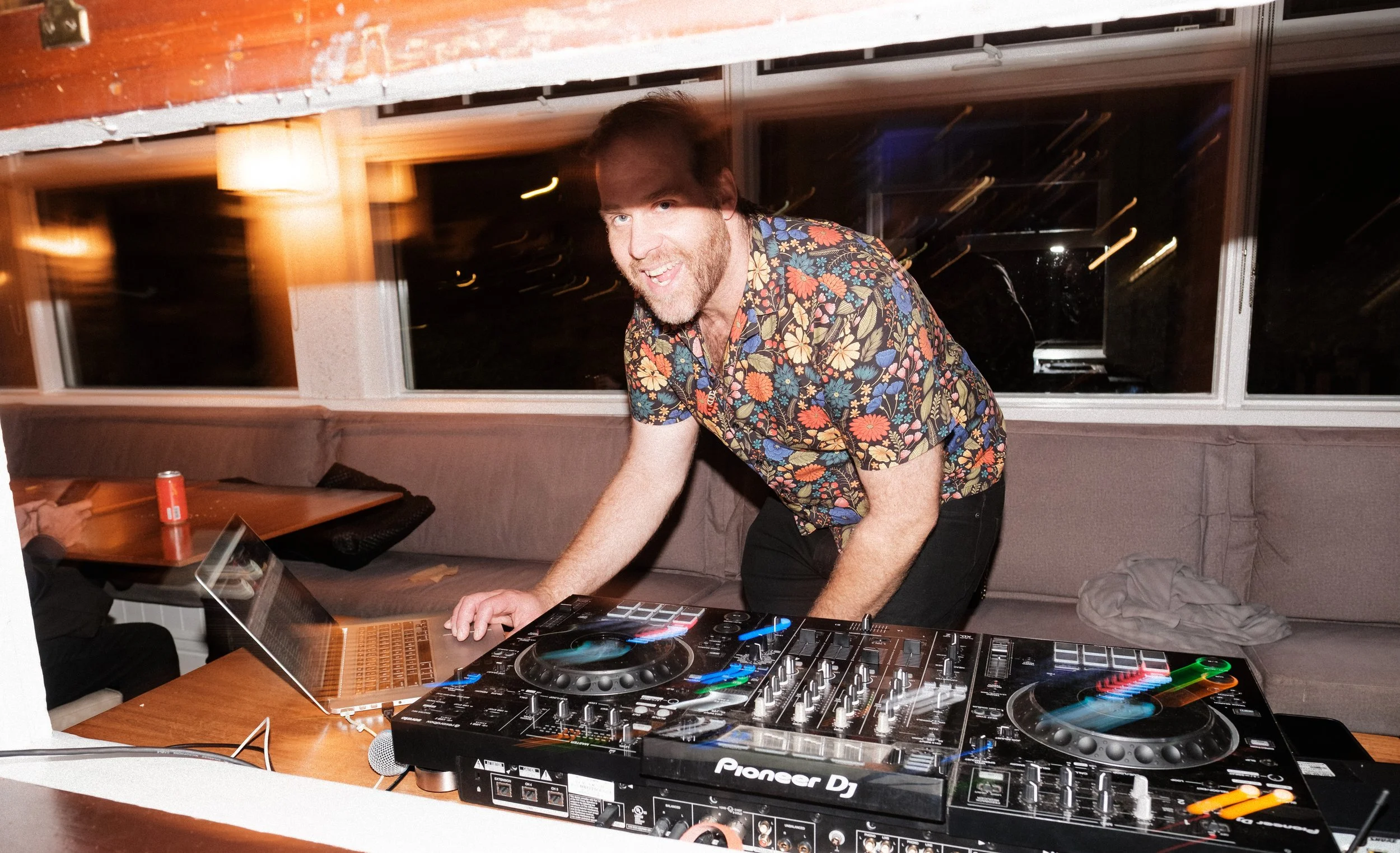 A man with brown hair and a beard, wearing a colorful floral shirt, is leaning over a Pioneer DJ setup and a laptop on a wooden table, smiling at the camera in a dimly lit room.