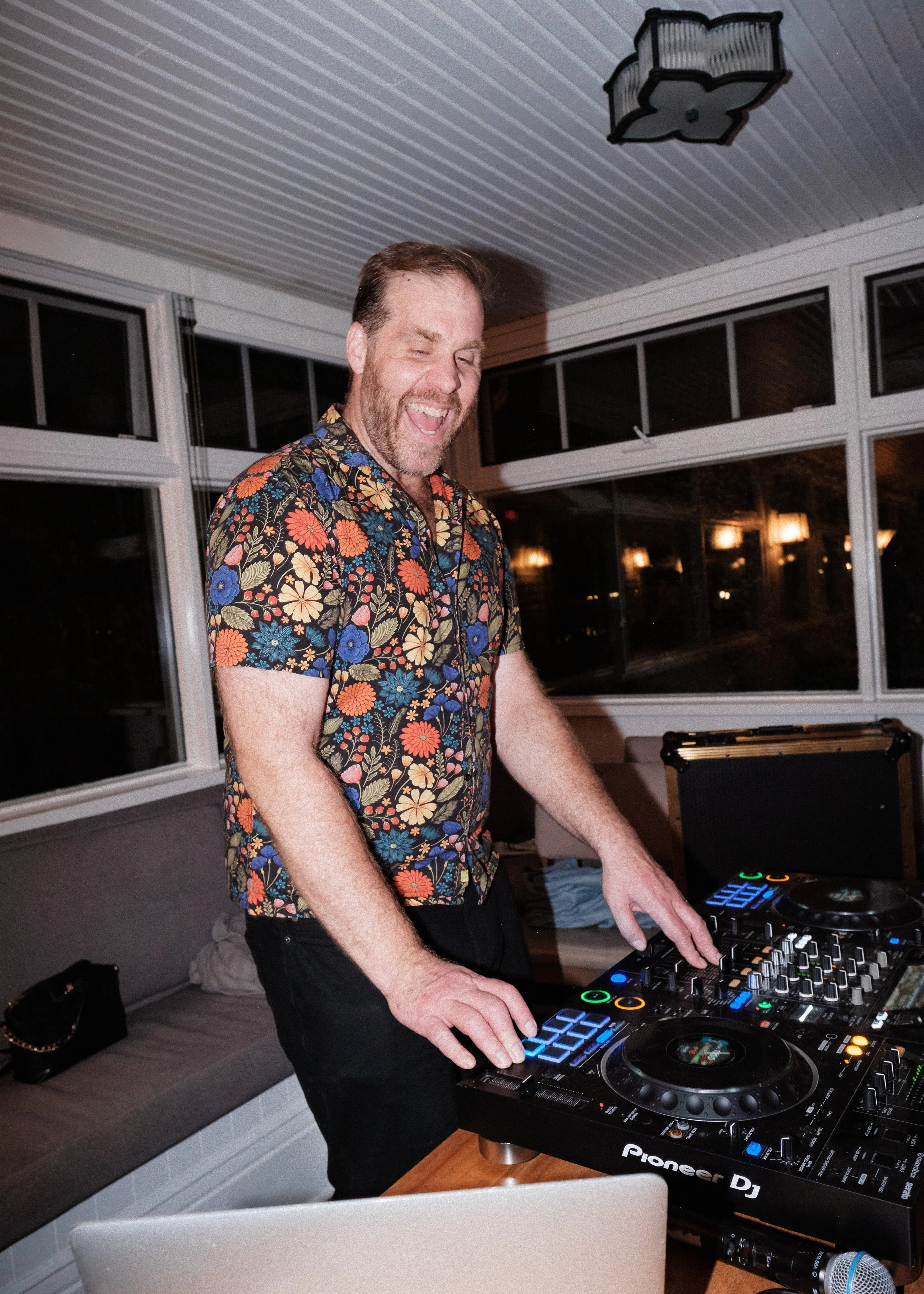A man with a beard and short hair wearing a colorful floral shirt, smiling and DJing with professional DJ equipment in a bright, modern room at night.