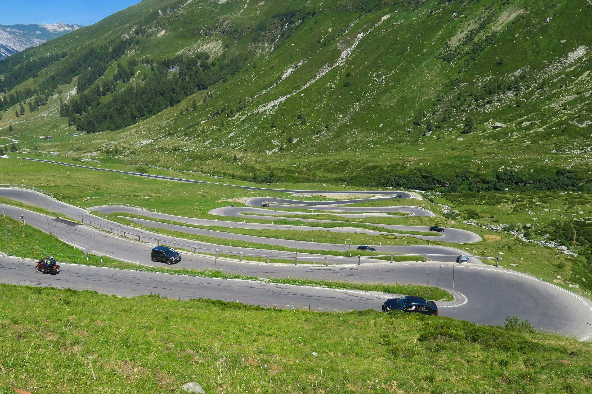 A mountain road with multiple switchbacks winding up a grassy hillside, with a few cars and a motorcycle riding along it, surrounded by green, mountainous terrain.