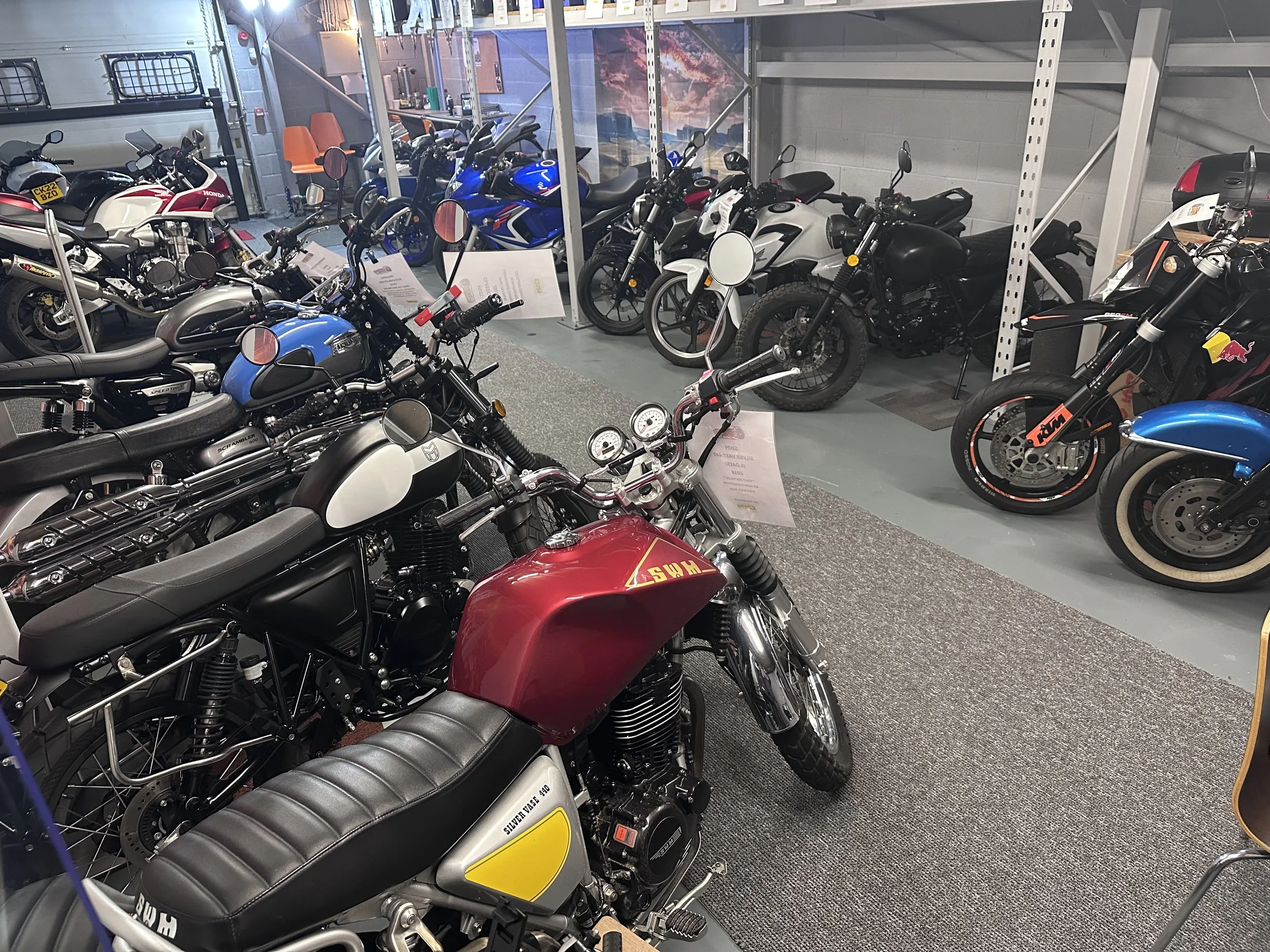 A showroom with multiple motorcycles on display, including a red SWM bike in the foreground and various other models lined up in the background, some with informational signs.