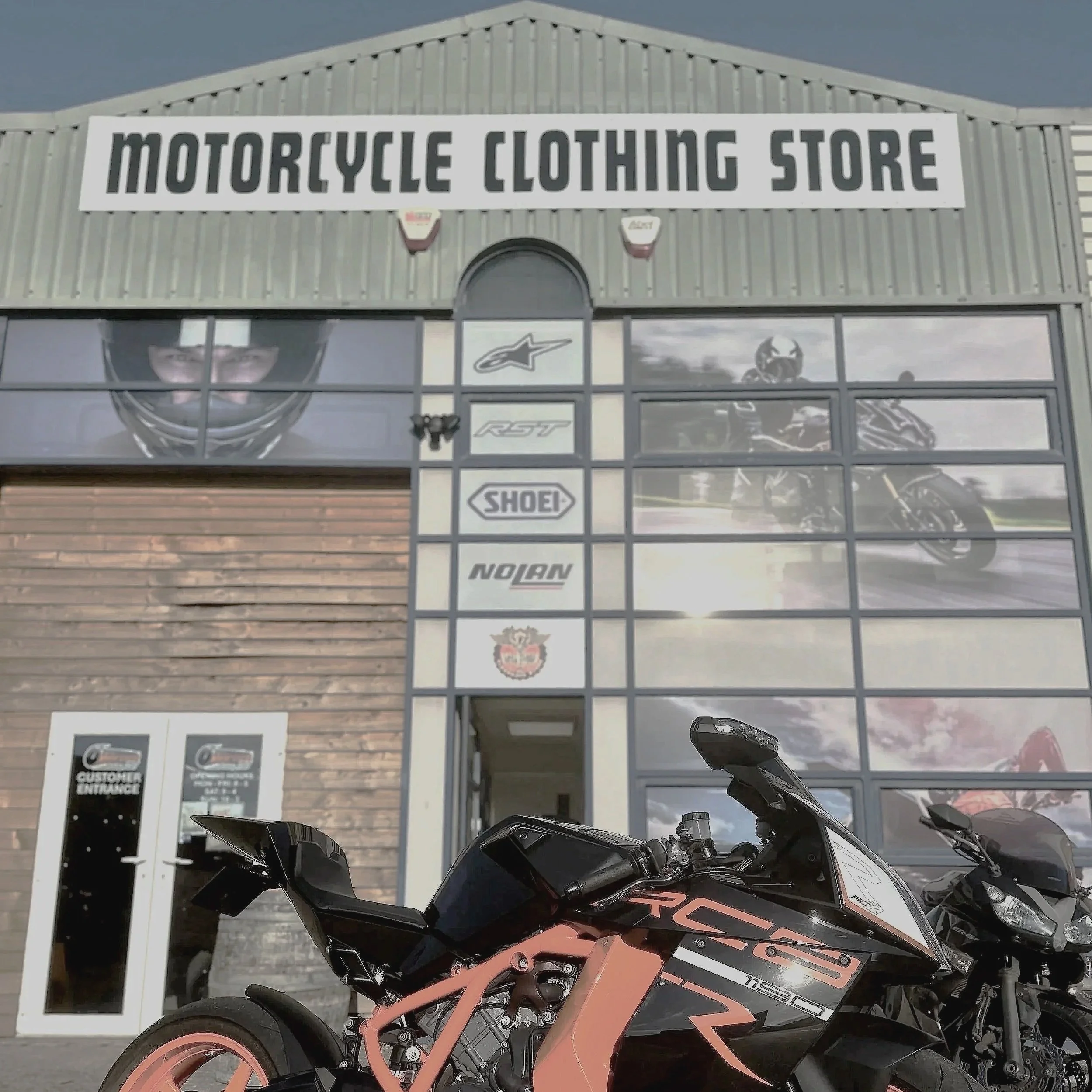 Exterior of a motorcycle clothing store with large glass windows showing motorcycle images and logos, a black and pink motorcycle parked outside, and an entrance door labeled 'Customer Entrance'.