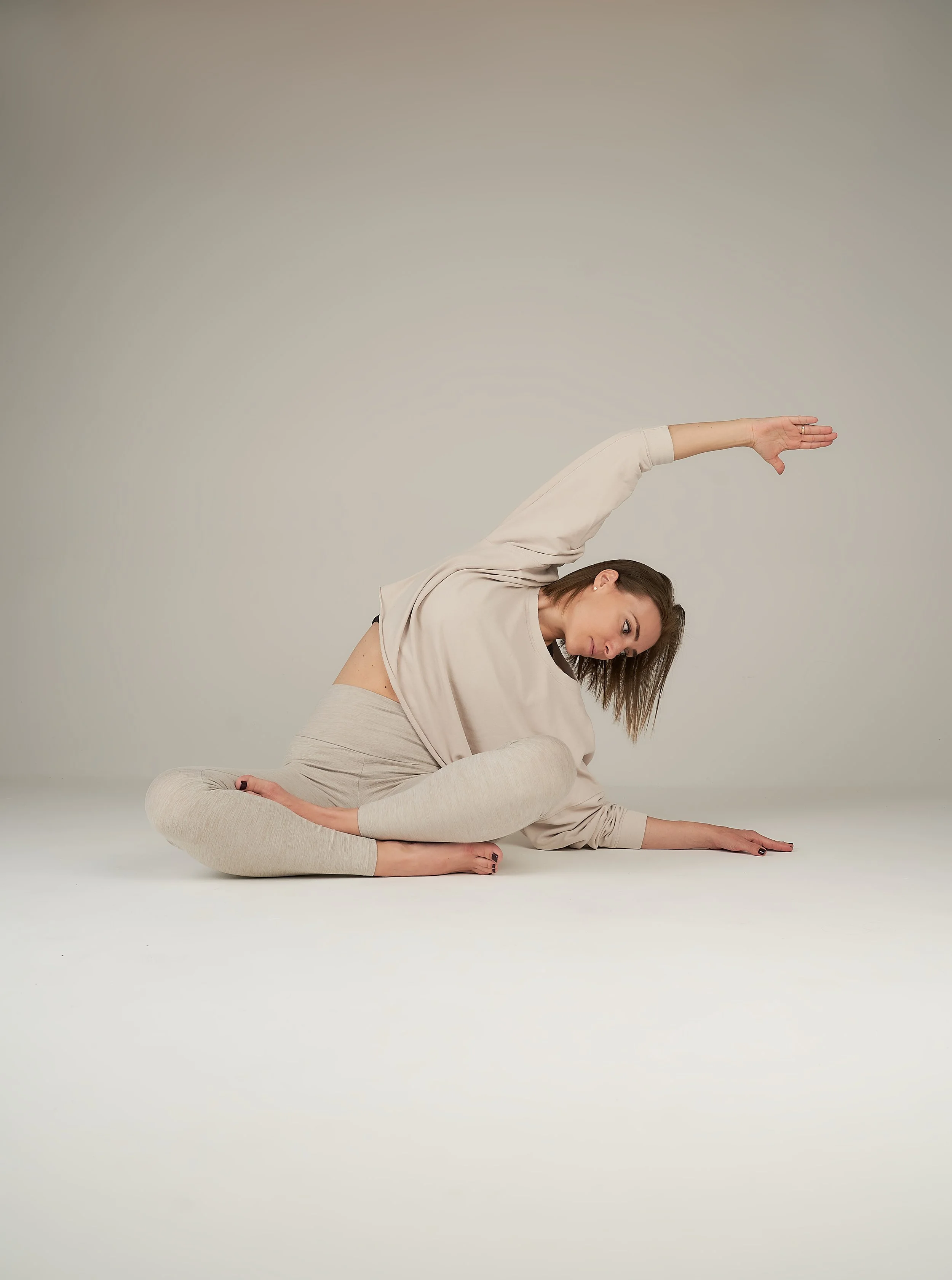 Woman in beige sweatshirt and light gray leggings doing a seated side bend yoga pose on a plain white floor against a light gray wall.