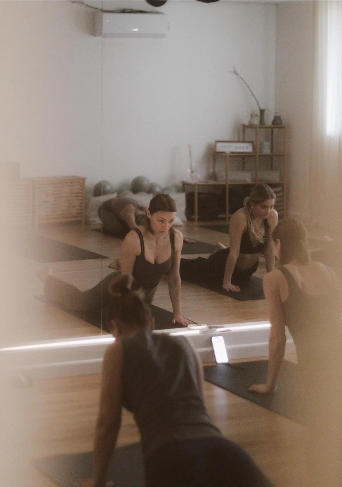 A woman practicing yoga in front of a large mirror in a fitness studio, with her reflection visible. She is on a black yoga mat, with a wooden floor and a shelf with decorative items in the background.