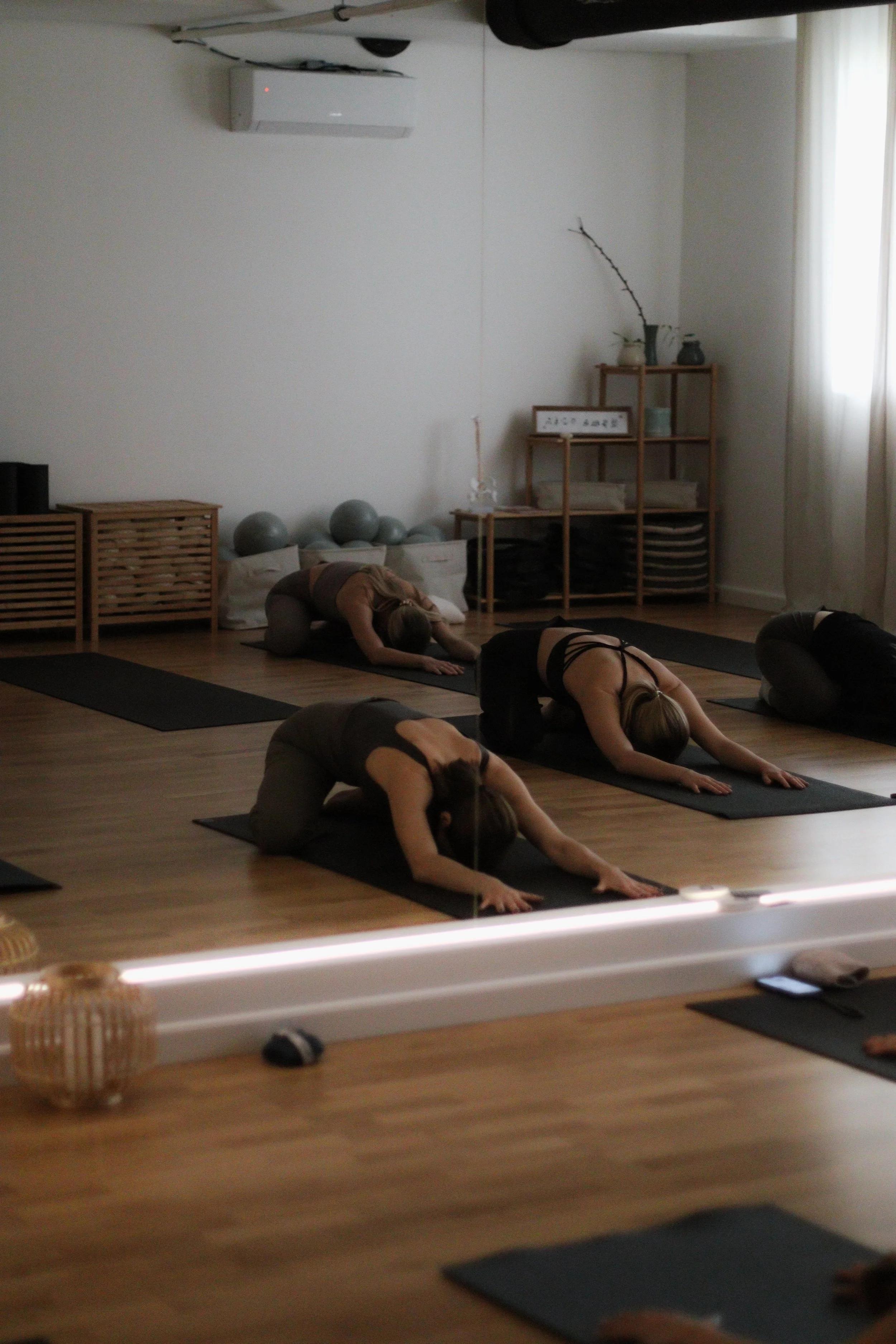 Women participating in a yoga class performing Child's Pose on yoga mats in a well-lit studio with wooden flooring, shelves, and decorative items.