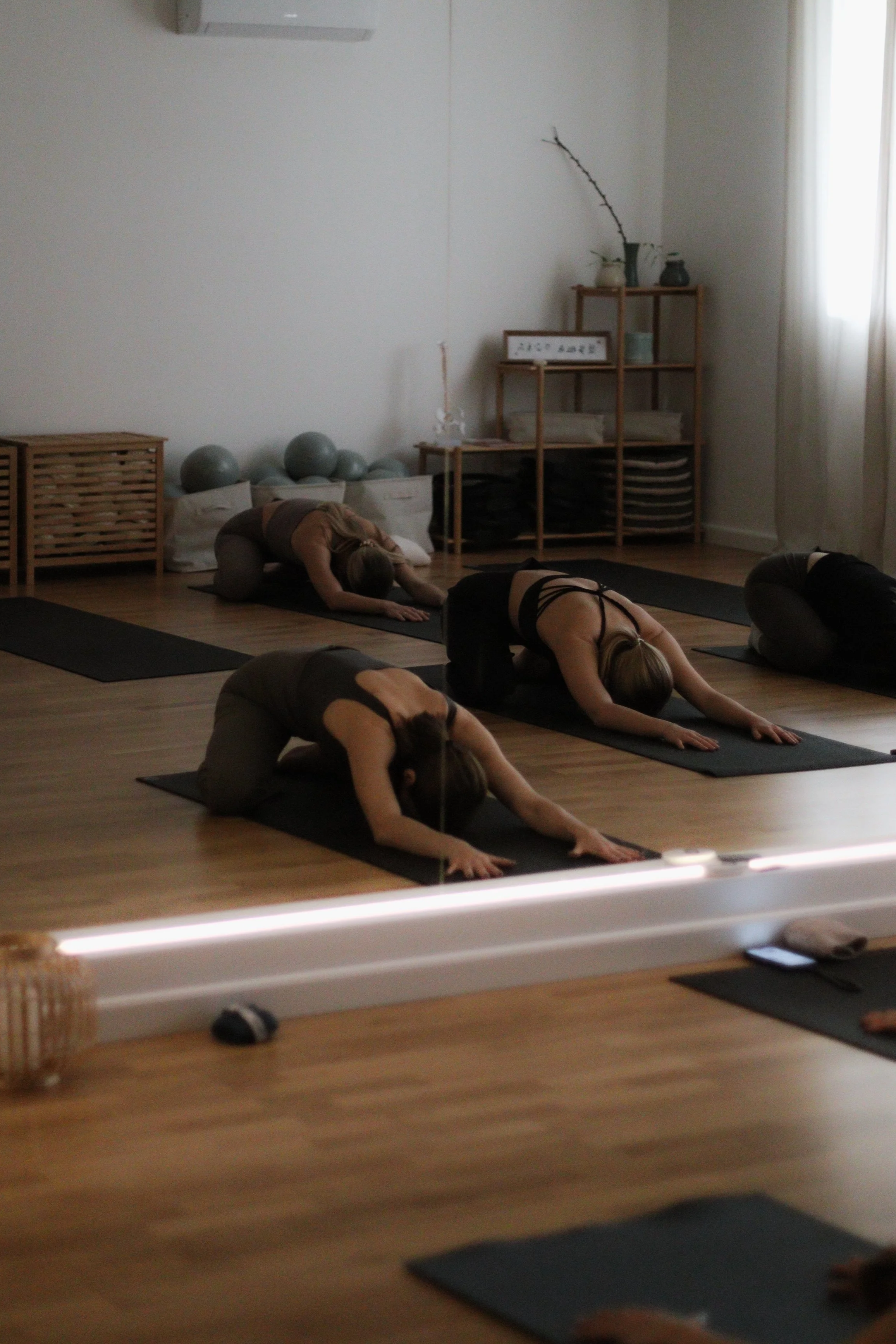 People practicing yoga in a dimly lit studio, positioned on black mats on a wooden floor.