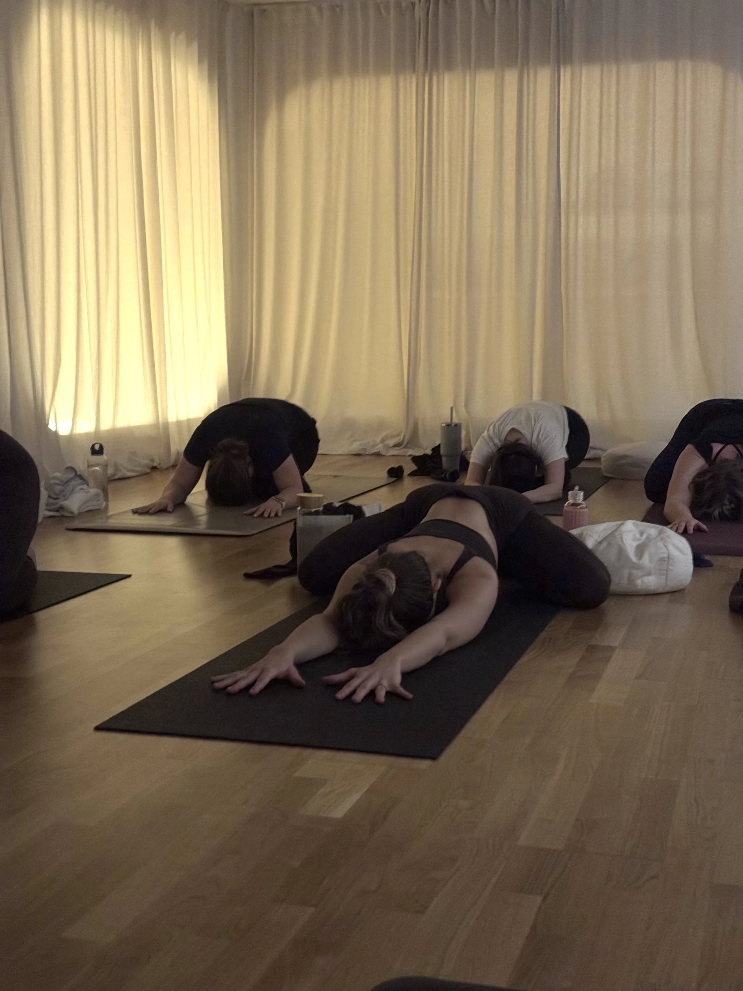 People participating in a yoga class, performing child's pose on yoga mats in a dimly lit room with cream curtains.