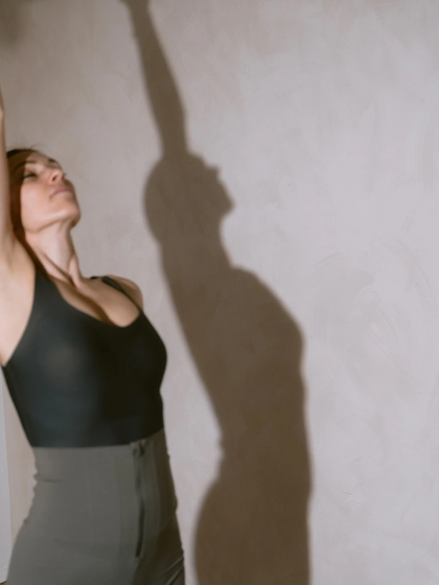 A woman with shoulder-length hair dressed in a black sleeveless top and gray pants, standing against a plain wall, casting a shadow on the wall.