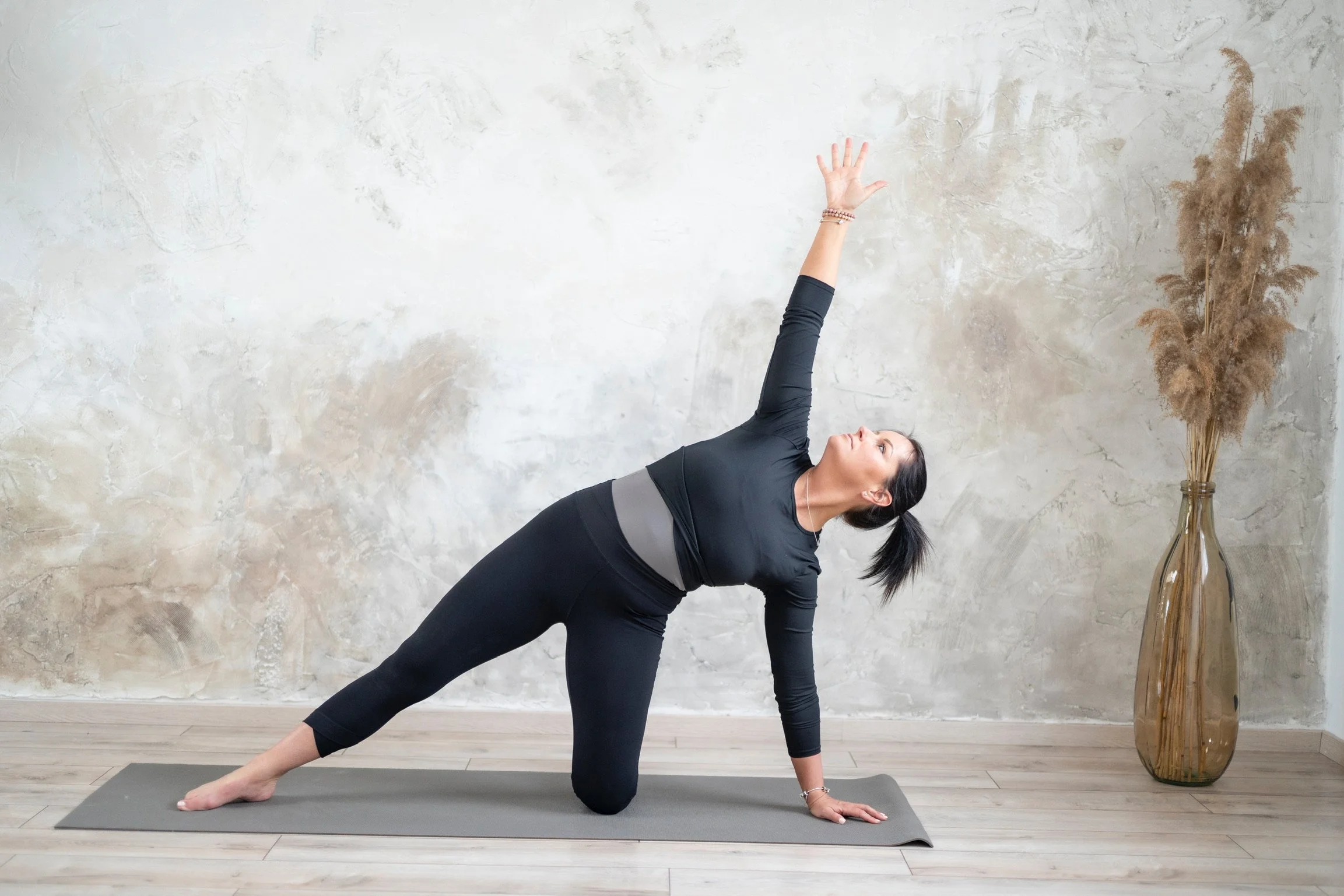 Woman practicing yoga in a variation of triangle pose on a gray mat in a minimalist room with a concrete wall and a tall vase with dried pampas grass.