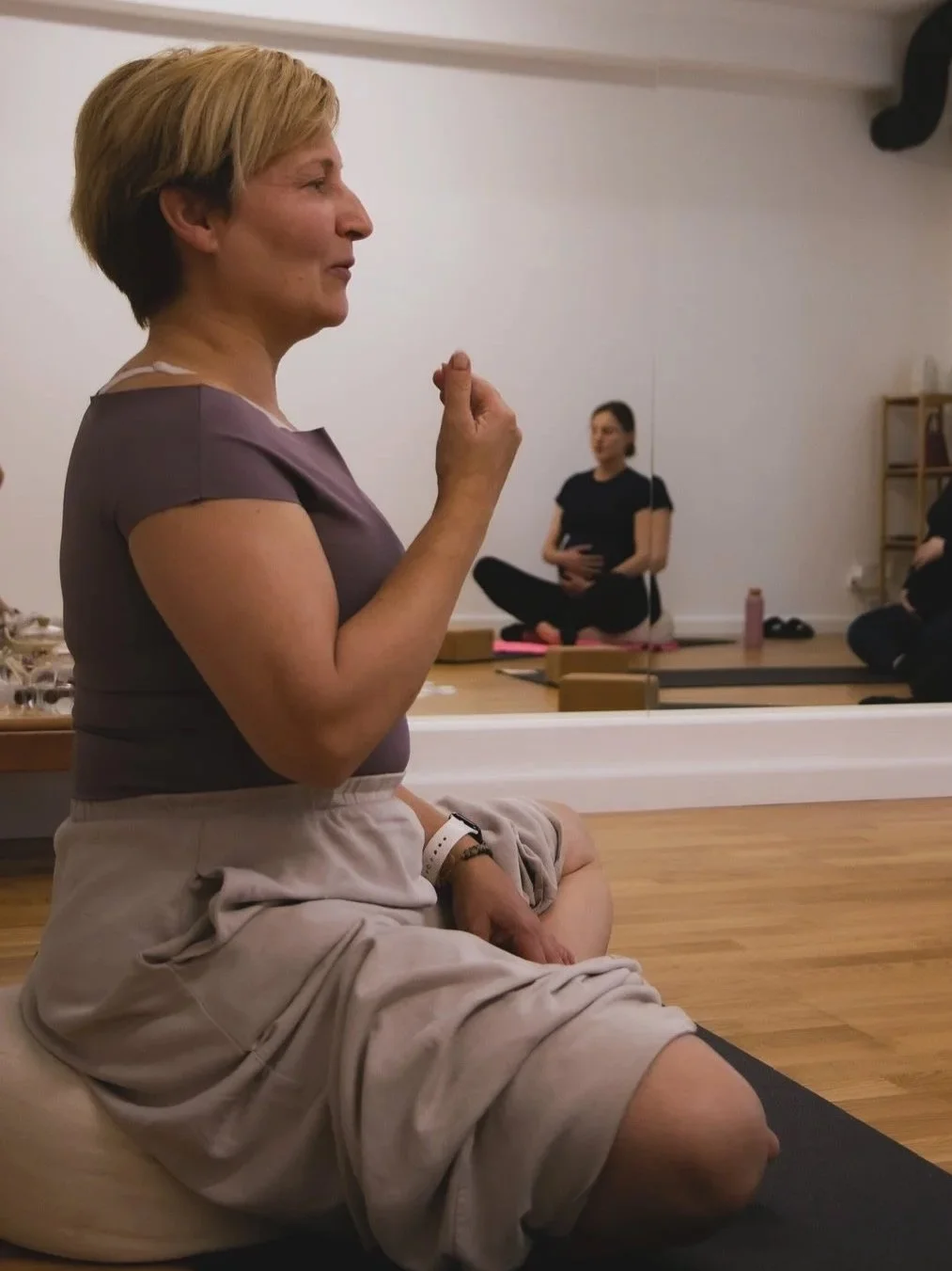 A woman sits cross-legged on a yoga mat in a meditation pose, with her hand in a mudra, in a yoga studio with a mirror reflecting other participants.