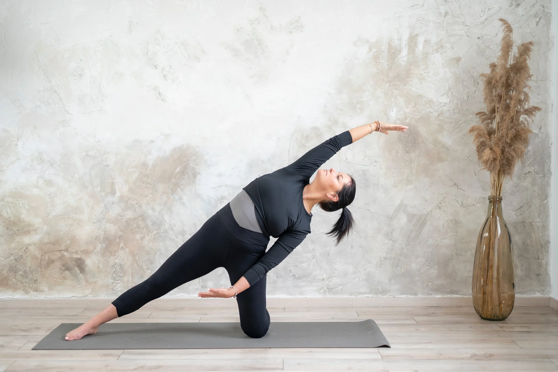Woman practicing yoga in a lunge twist pose on a yoga mat in a room with a textured beige wall and a tall vase with pampas grass.