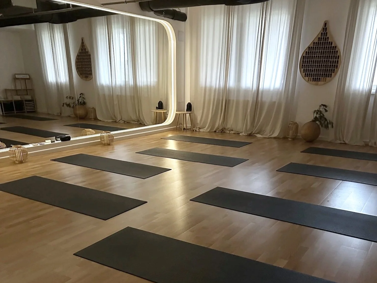 A yoga or meditation studio with black mats on a wooden floor, a large mirror, and minimalist decor including plants and decorative woven items.