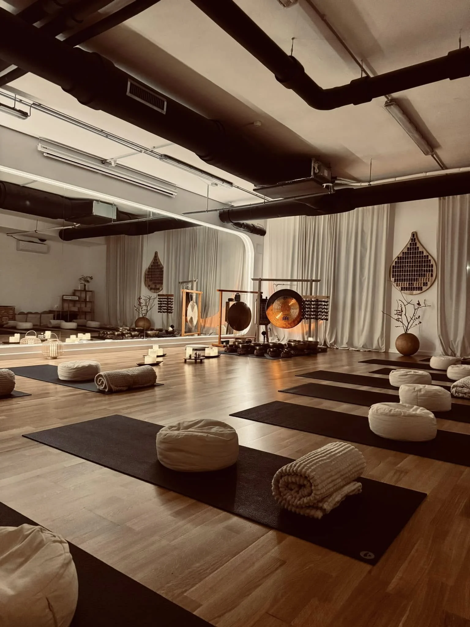 Yoga mats, bolsters, and rolled towels arranged on a wooden floor in a dimly lit studio with hanging gongs, candles, and decorative wall hangings, creating a serene atmosphere.