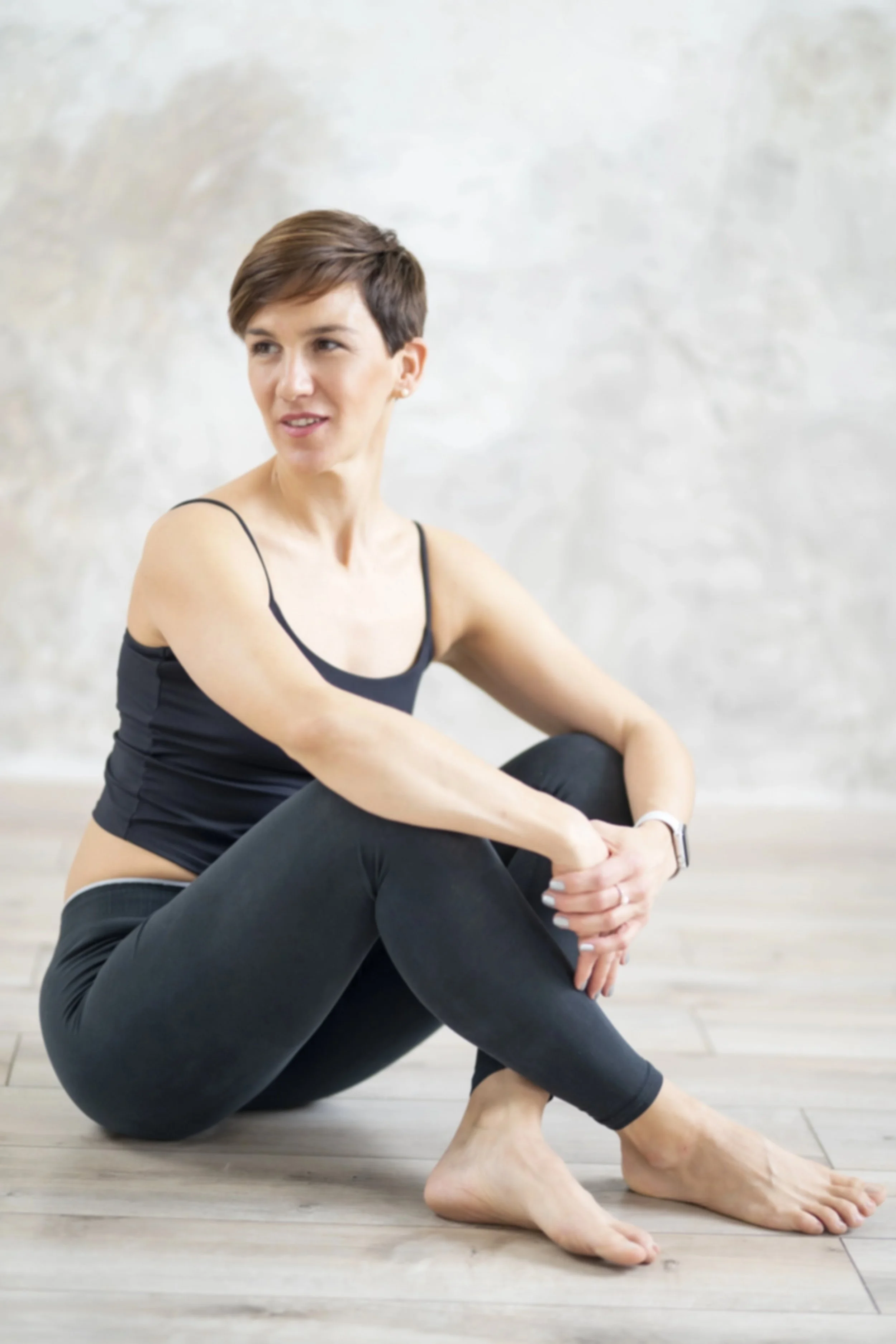 Woman with short brown hair sitting on the floor, wearing a black tank top and black leggings, with a neutral background.