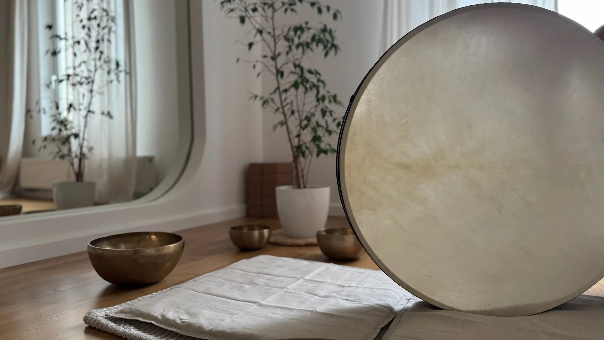 A round cymbal, three brass singing bowls, a white cloth, and a large mirror on a wooden floor in a minimally decorated room with a potted plant and a window with sheer curtains.