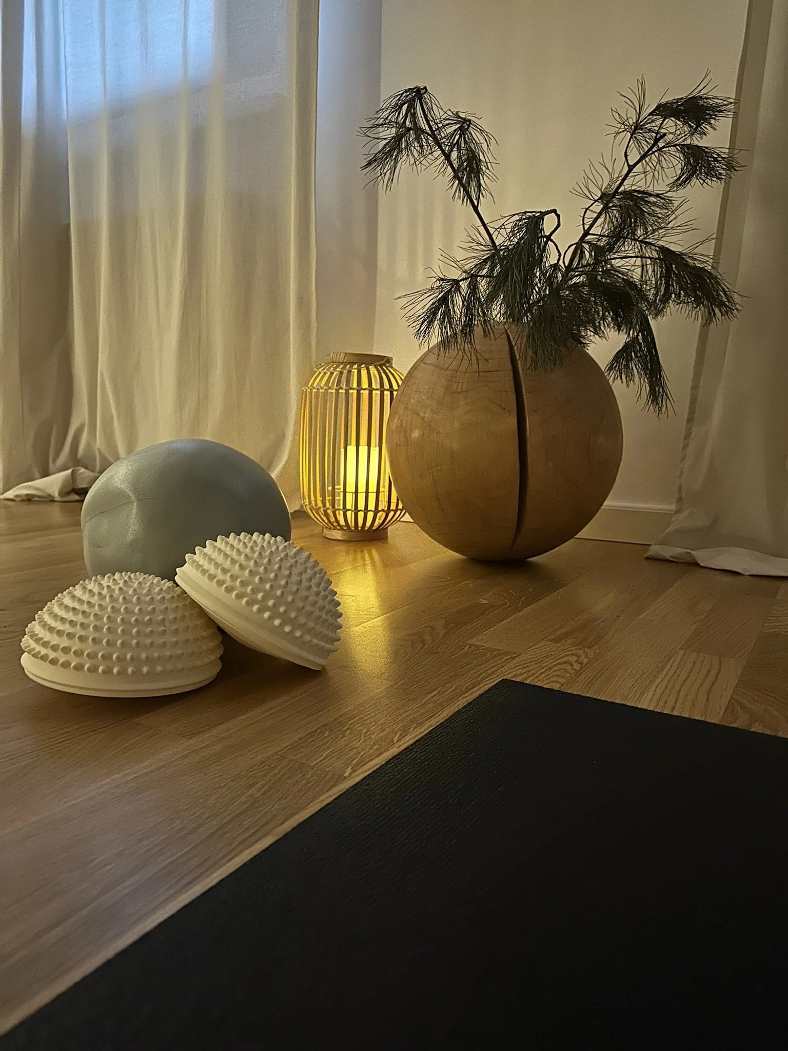 A wooden table with decorative items including a large wooden vase with dried branches, a small yellow lantern, a gray sphere, and two white textured sculptures, with curtains in the background and soft lighting.