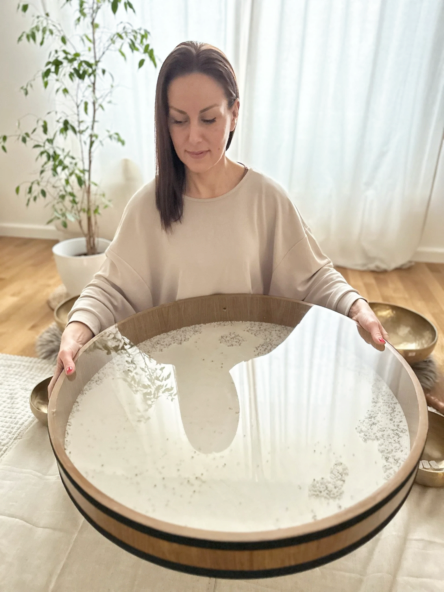 A woman with brown hair looking down, holding a large wooden tray with a mirror-like surface, surrounded by a cozy room with plants and decor.