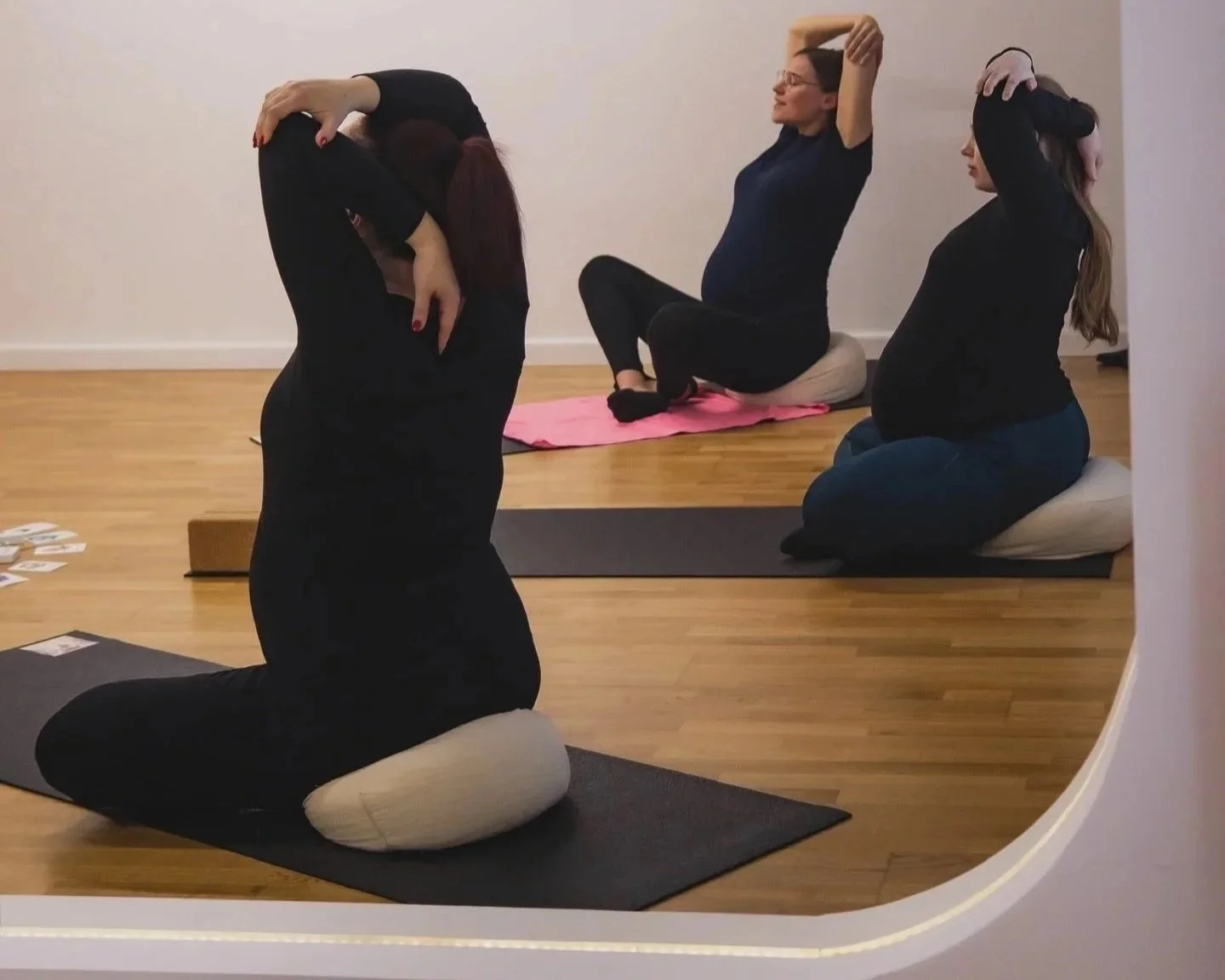 Three women practicing yoga in a room with wooden floors, using cushions and mats. One woman is kneeling with a forward bend, the other two are sitting on kneeling cushions, each with one arm raised and their hands behind their heads.