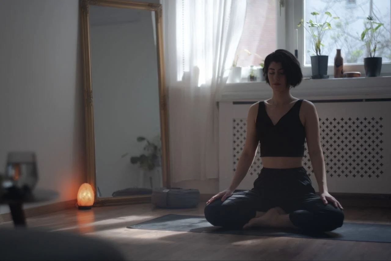 A woman practicing yoga in a peaceful, well-lit room, sitting cross-legged on a yoga mat, with a mirror and houseplants in the background.