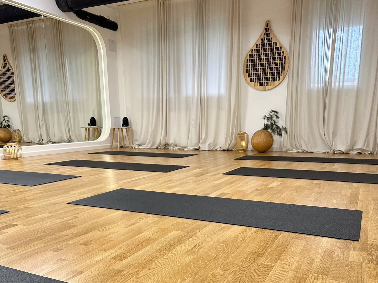 Yoga studio with black mats on a wooden floor, large mirror, beige curtains, wall decorations, and potted plants.