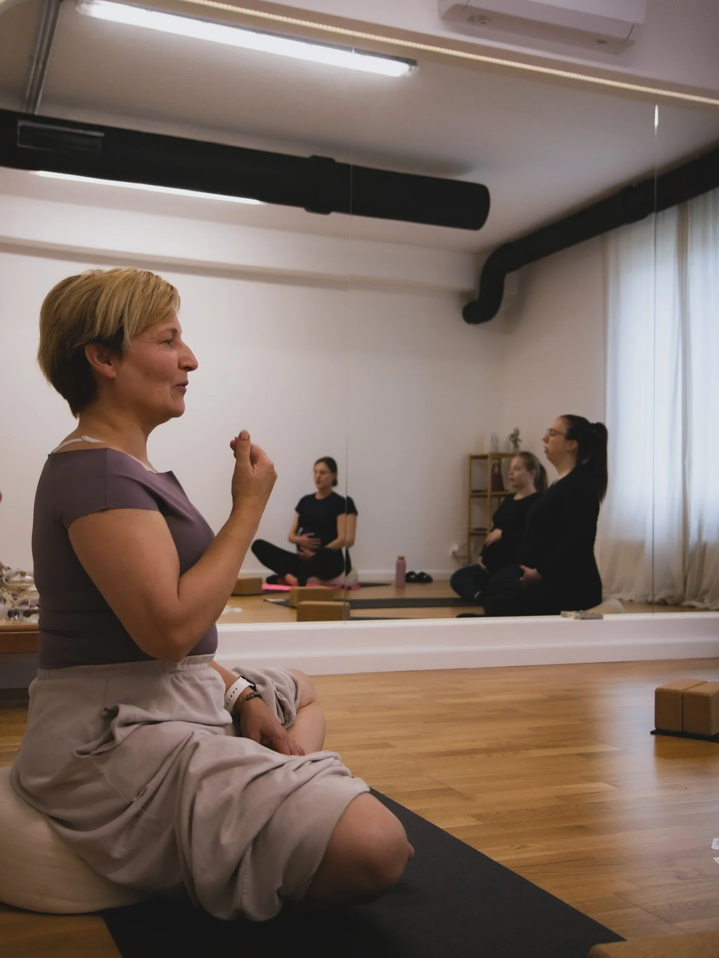 A group of women practicing meditation or yoga in a mirrored studio with wooden floors, some sitting cross-legged on yoga mats, and one woman in the foreground sitting on a cushion with closed eyes, wearing a purple top and beige shorts.