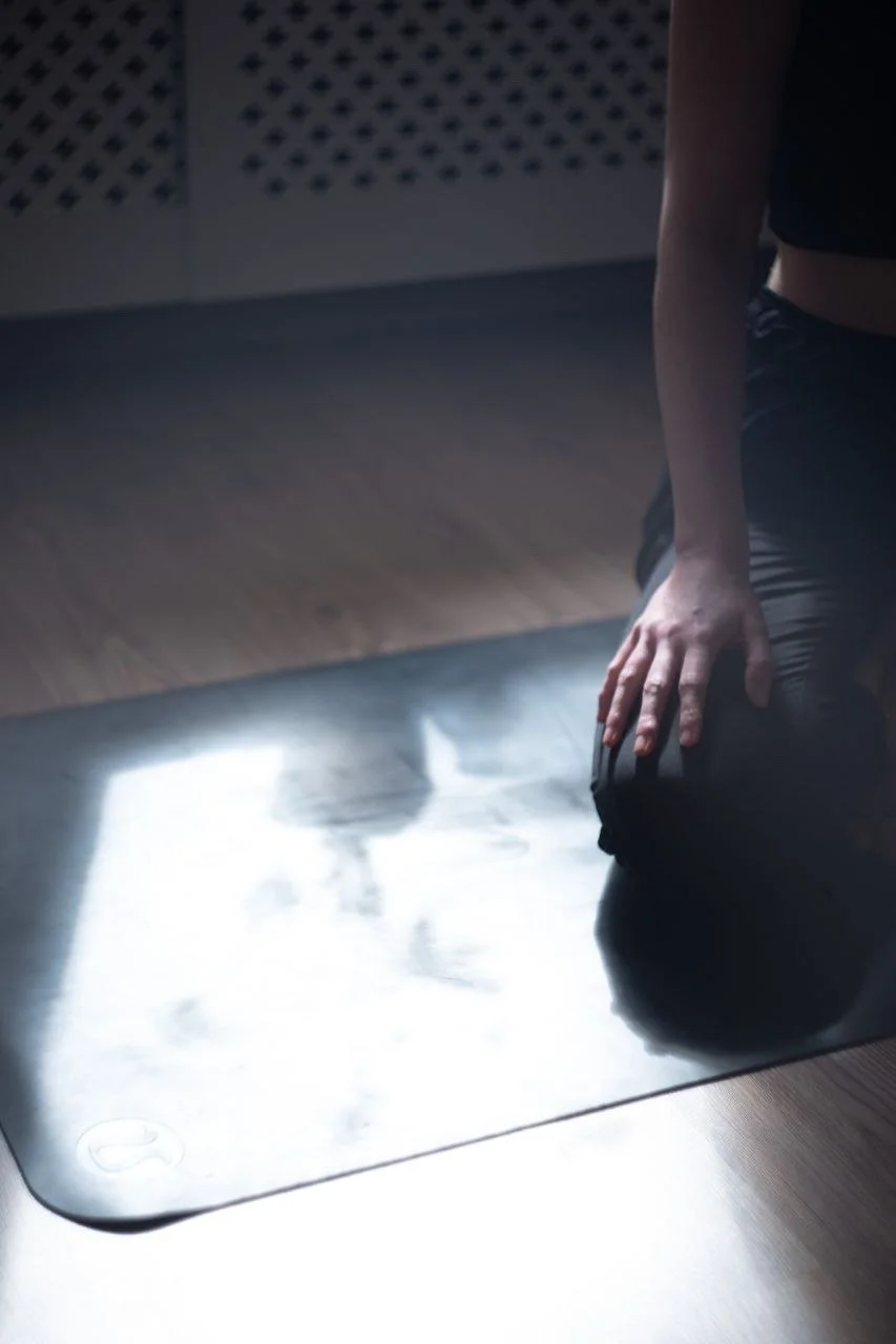 Person kneeling on a black yoga mat, touching their thigh, in a dimly lit room with a reflective surface.