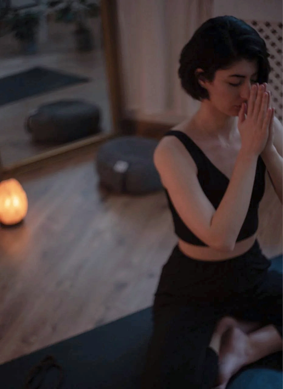 A woman with short black hair practicing yoga or meditation, sitting cross-legged on a yoga mat with her hands pressed together in front of her face, in a dimly lit room with a candle and mirror in the background.