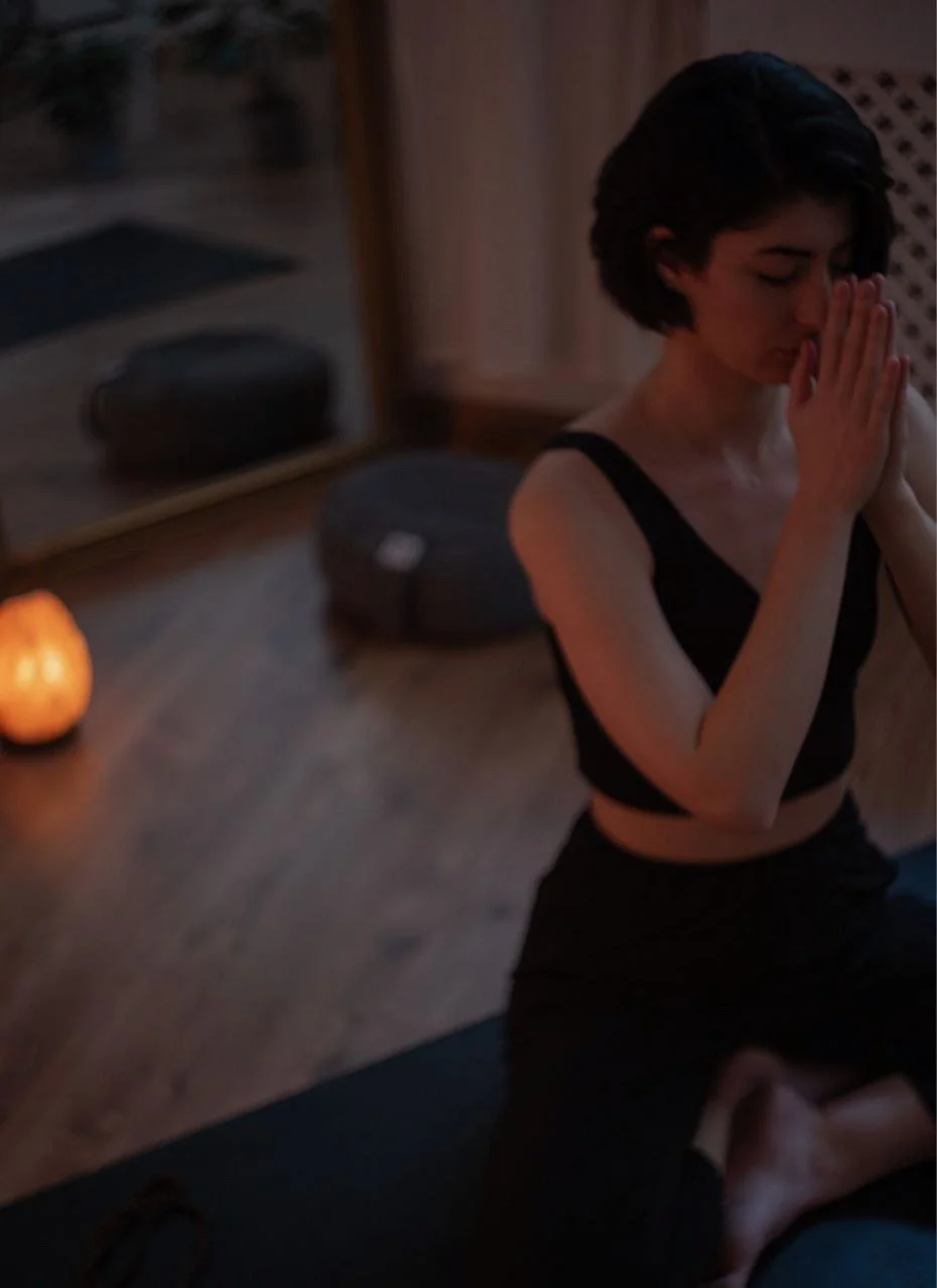 A woman practicing yoga or meditation in a dimly lit room, sitting cross-legged on a yoga mat with hands in prayer position, with a small glowing lamp and a speaker visible in the background.
