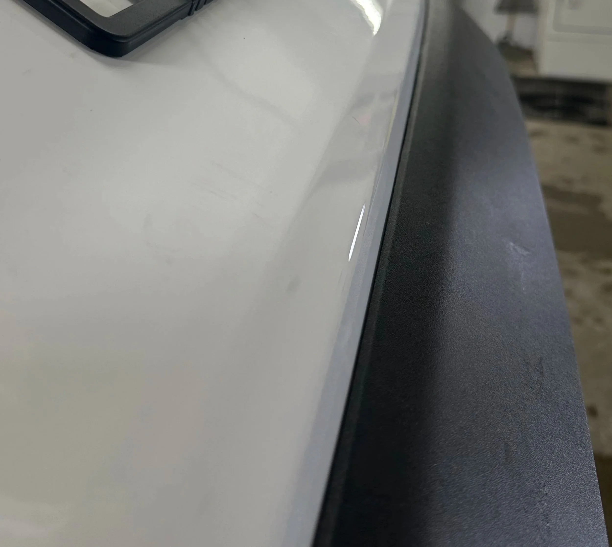 Close-up of a white vehicle's hood with a black badge that reads 'VANHOUTEN,' and part of an indoor garage with a broom and laundry appliances in the background.