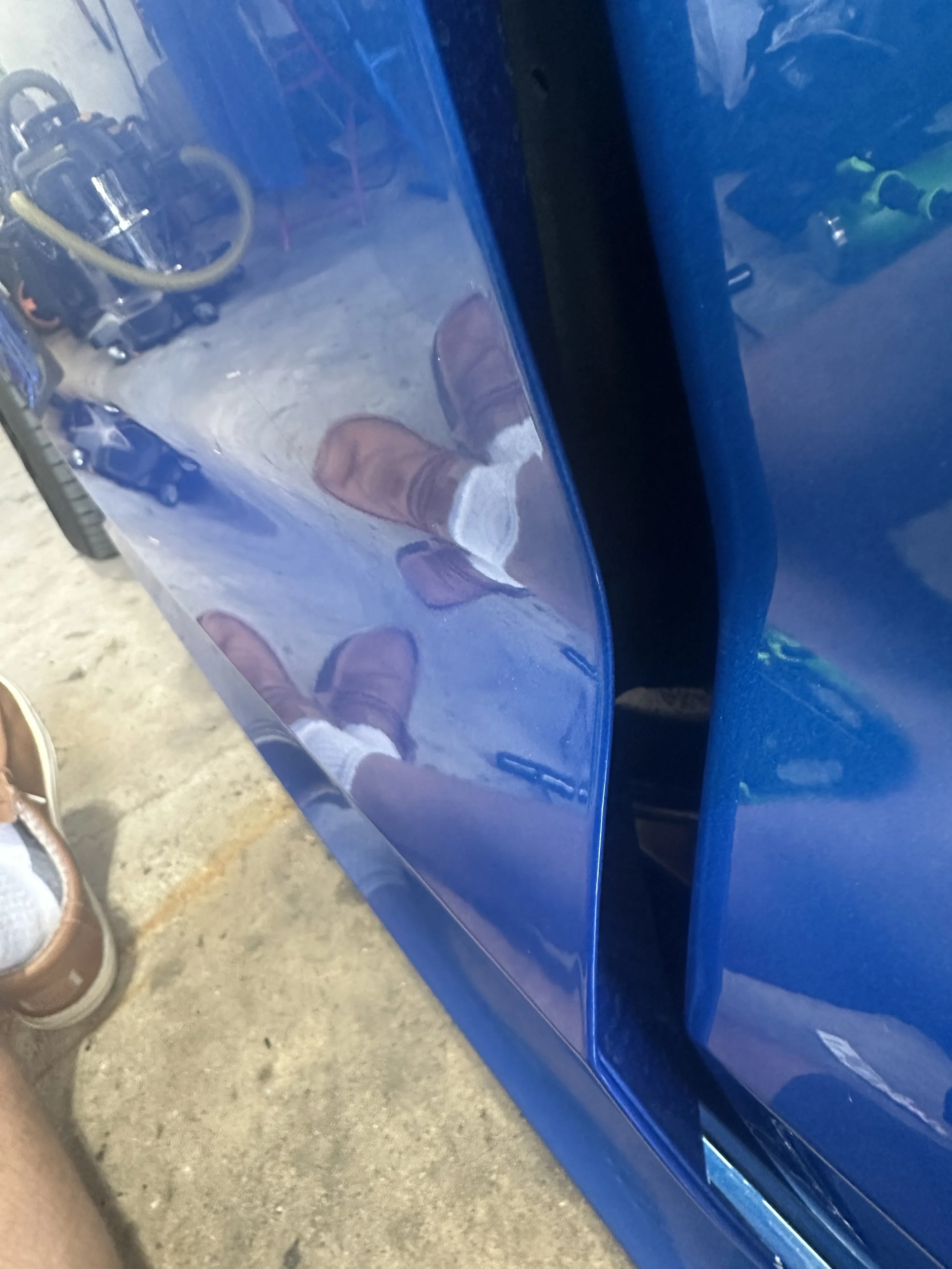 Reflection of a person wearing brown shoes, white socks, and dark shorts, standing on a concrete surface beside a shiny blue car.