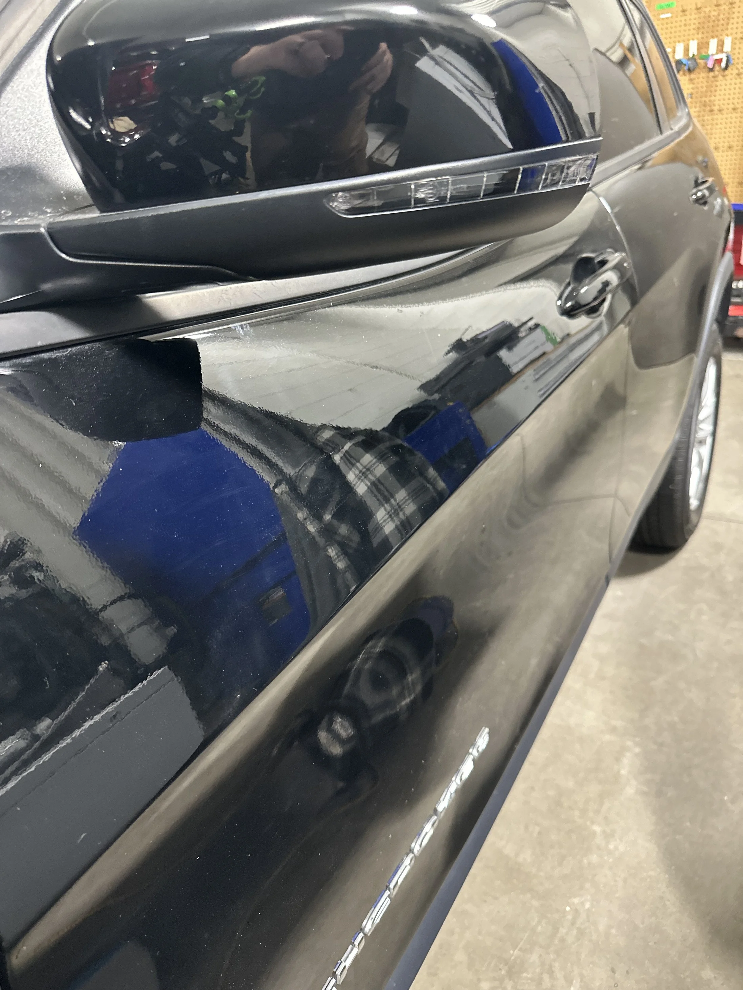 Close-up of a black car's side mirror and door, with reflections of a person taking the photo and the garage interior, including tools on a pegboard.