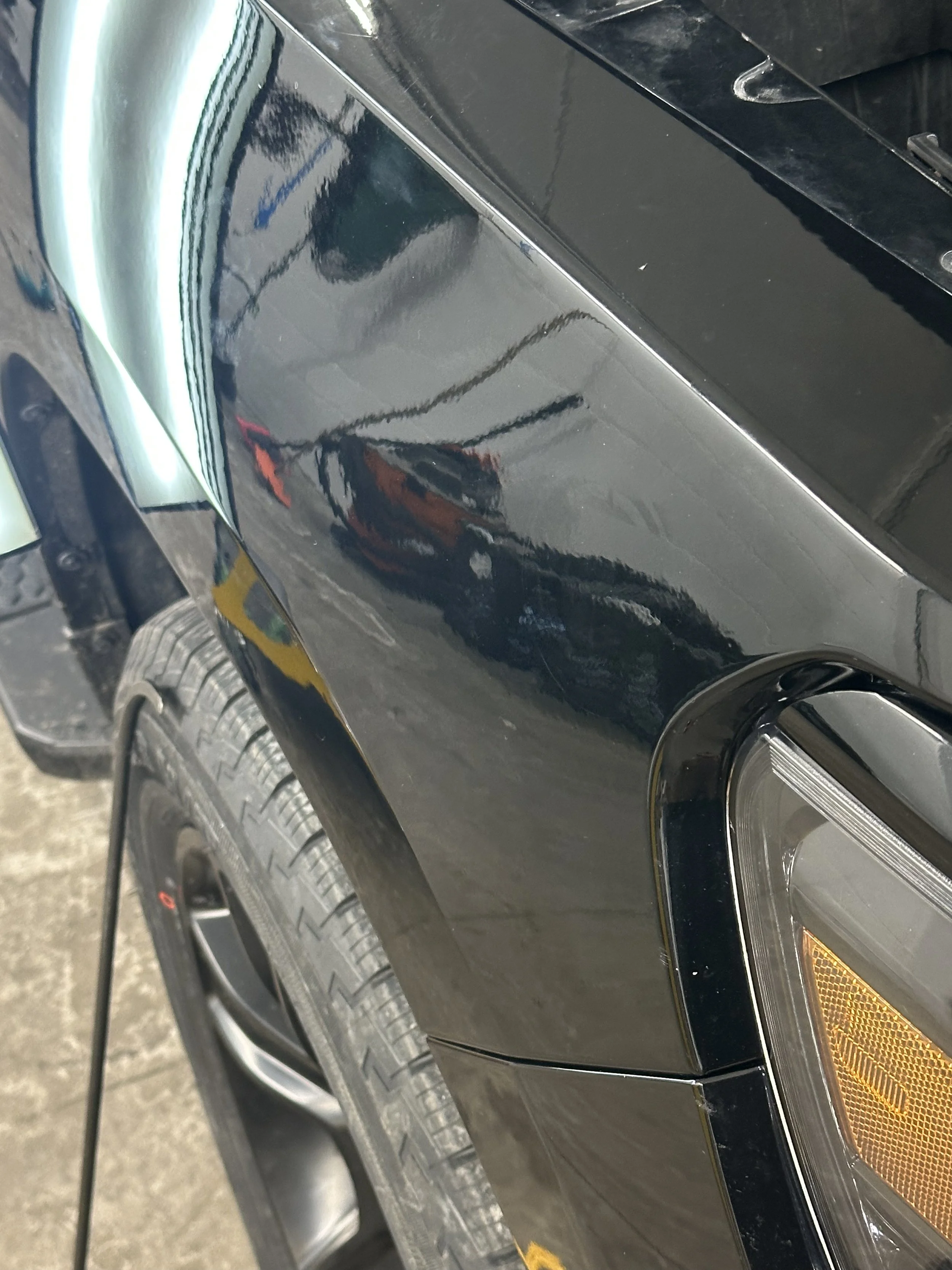 Close-up of a black car's front quarter panel, showing a noticeable scratch and slight dent near the wheel well, with a reflection of the garage interior and a person taking the photo.