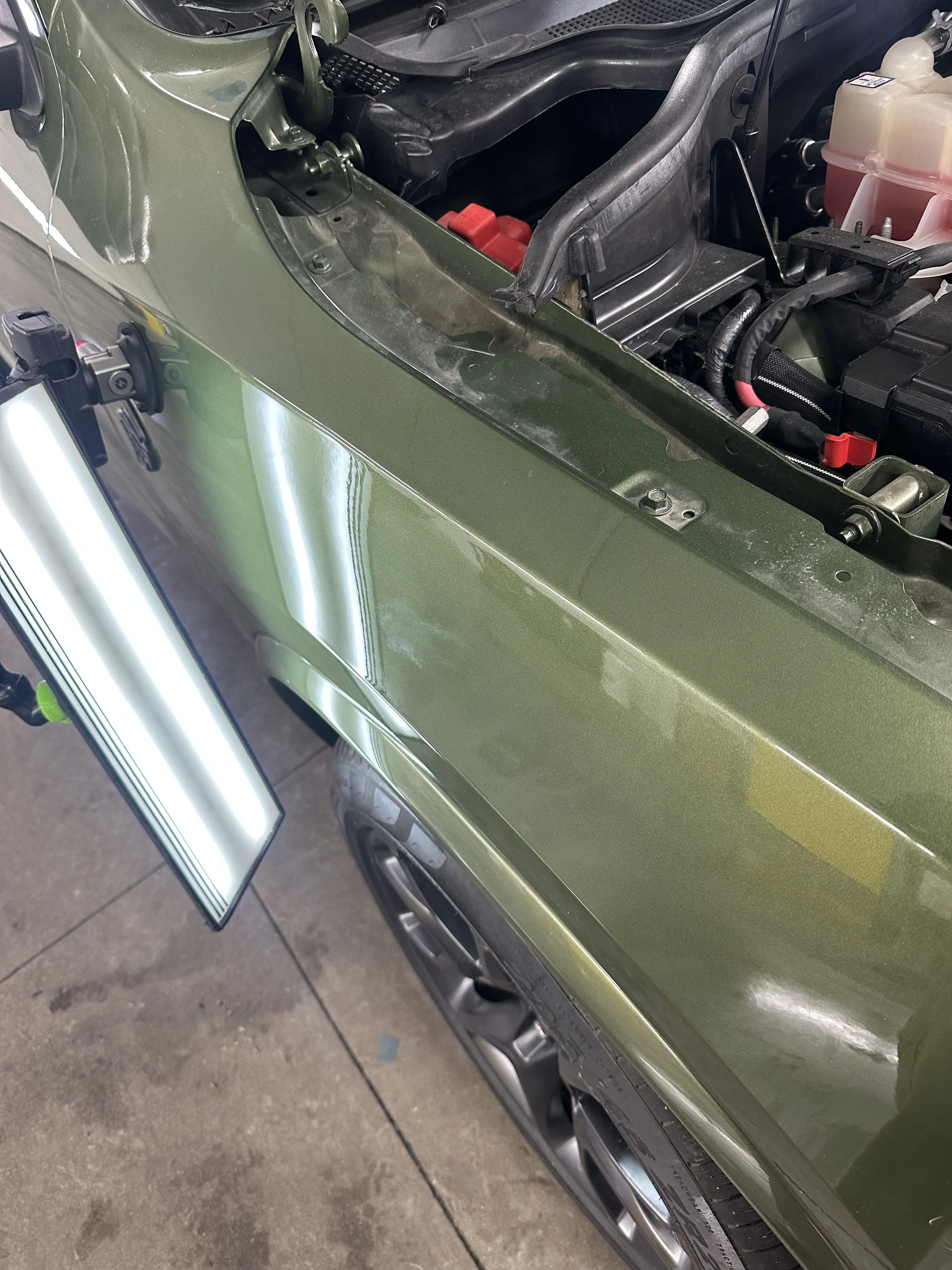 Close-up of a green car with its hood open, showing the engine compartment, fender, and wheel.