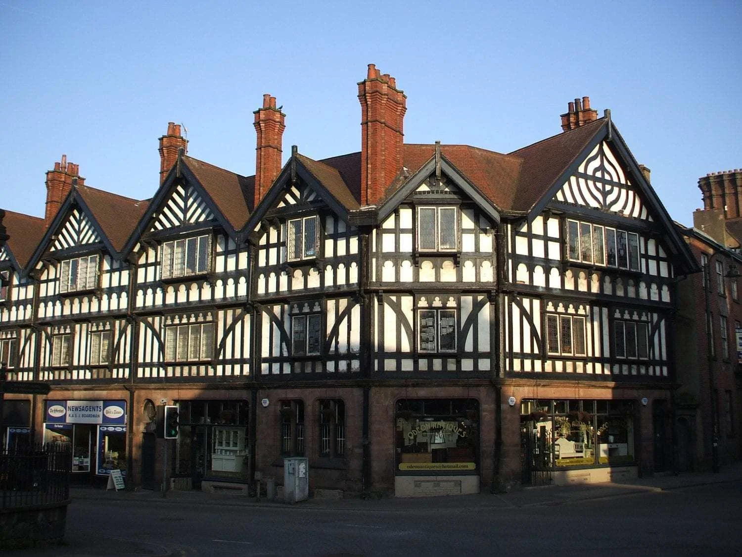 A historic building with black-and-white timber framing, tall brick chimneys, and multiple gabled roofs, located on a city street with a shopfront at the ground level.