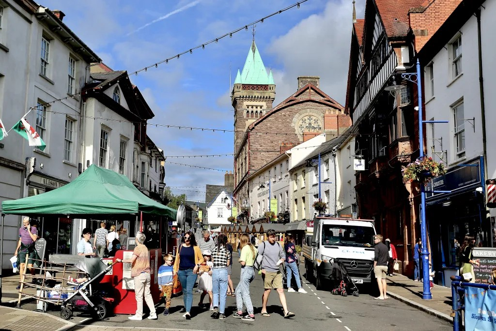 Busy street scene with pedestrians, market stall with a green canopy, historic buildings, and a church with a tall tower and clock in the background.