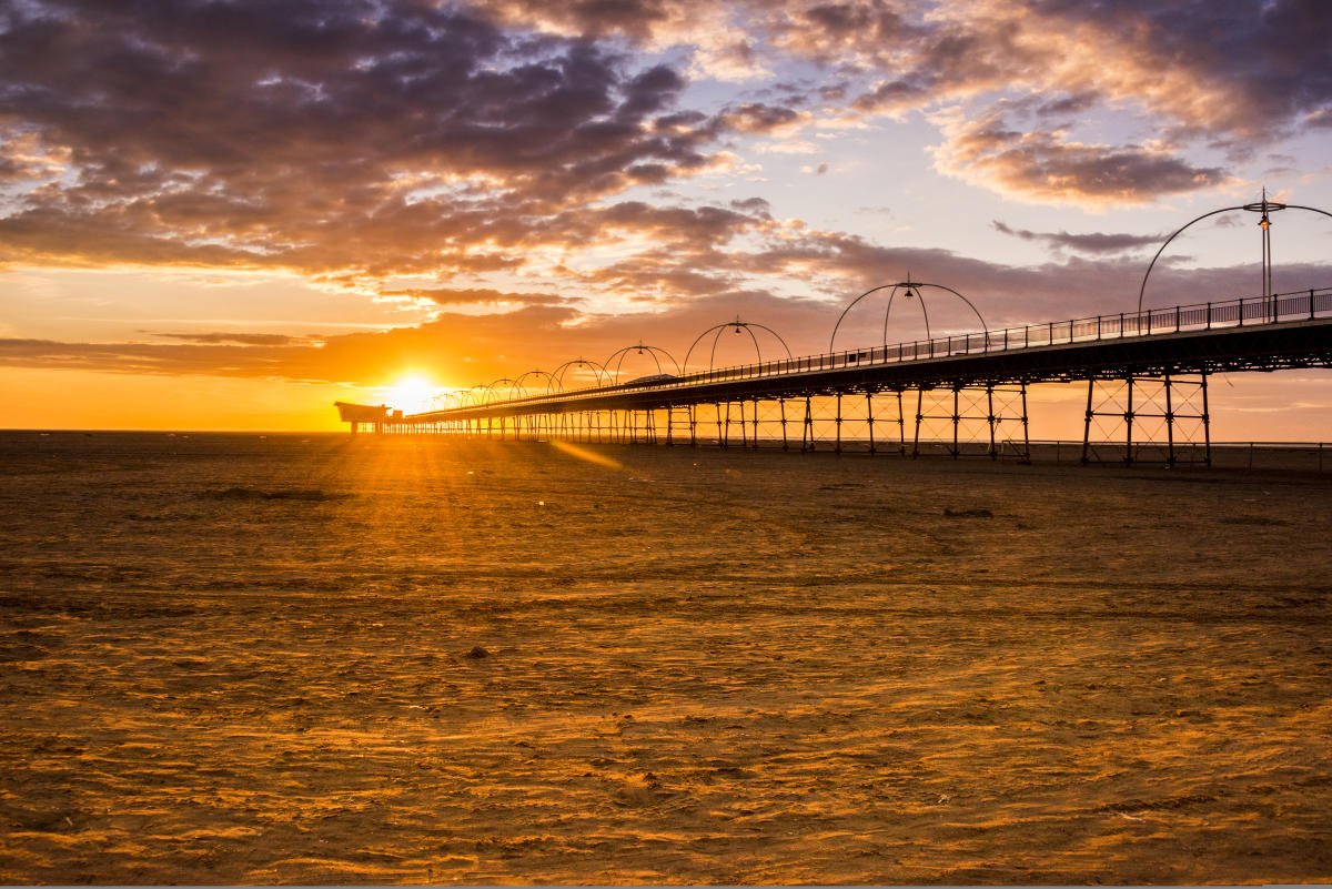 A pier extending into the ocean during sunset, with colorful clouds in the sky and the sun near the horizon.