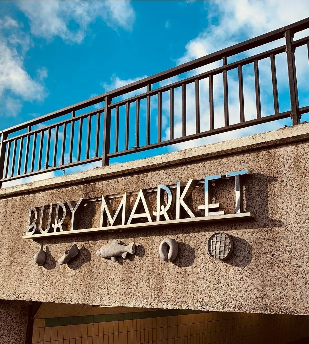 Sign reading 'Bury Market' with fish, seafood, and waffle-shaped decorations on a concrete wall beneath a blue sky with clouds.