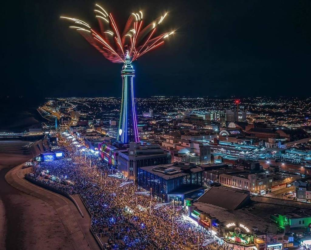 Nighttime city scene with the Space Needle illuminated in neon lights, fireworks exploding above it, and a large crowd gathered along the waterfront.