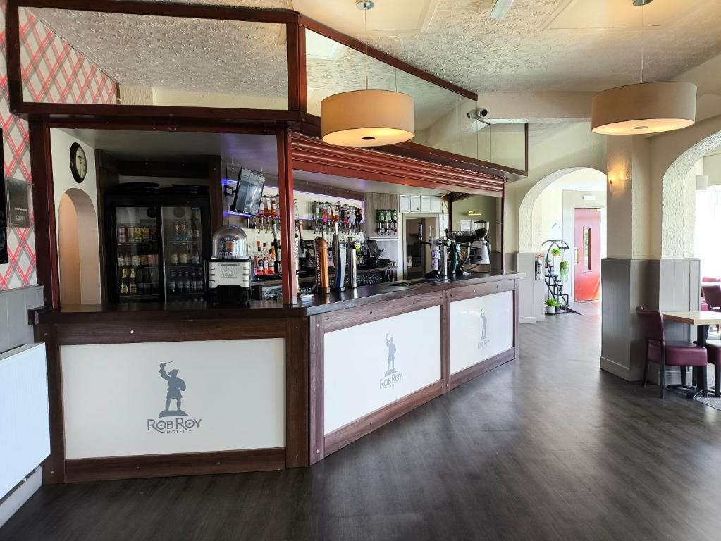 Empty hotel bar with dark wood counter, coffee machine, liquor bottles, soda dispenser, and a small refrigerator, in a well-lit space with hanging circular ceiling lights and a seating area nearby.