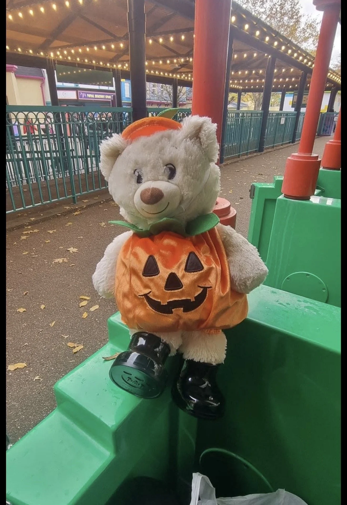 A teddy bear dressed in a Halloween costume resembling a pumpkin, with a carved face, green collar, and black shiny shoes, sitting on a green plastic bench at an amusement park during dusk, with string lights overhead and a decorative wooden structure in the background.