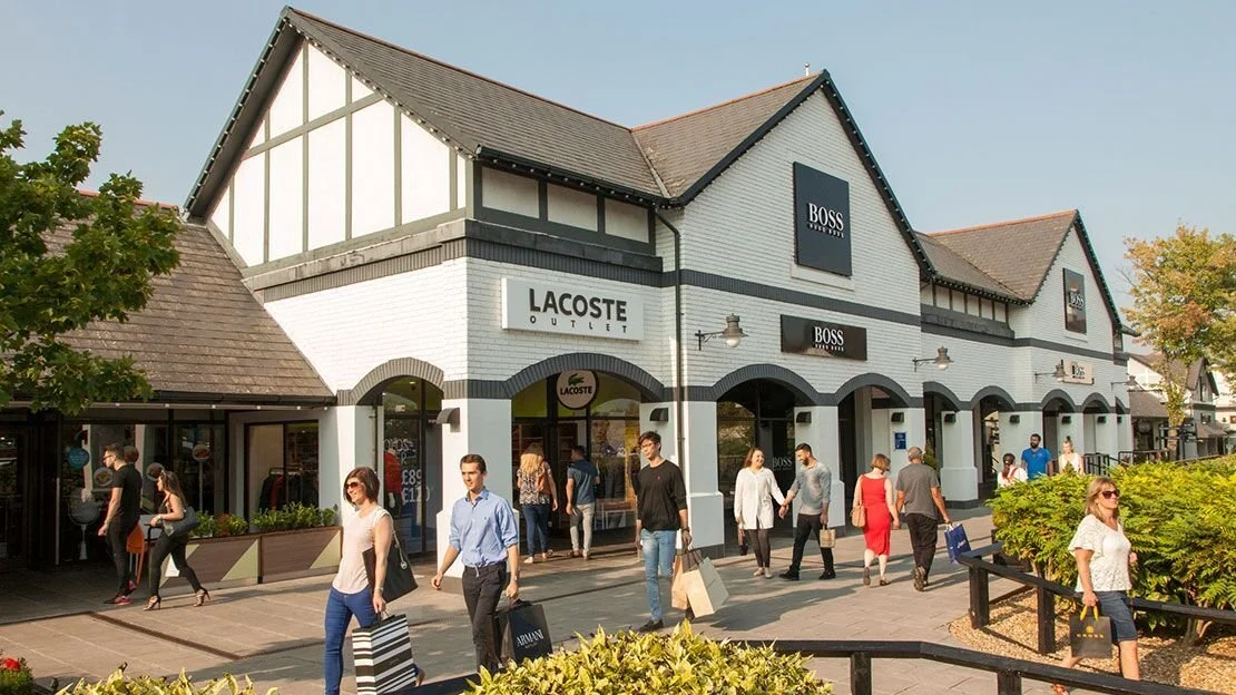 People walking outside a shopping outlet with stores like Lacoste and Boss, in front of a building with a gabled roof and white brick walls.