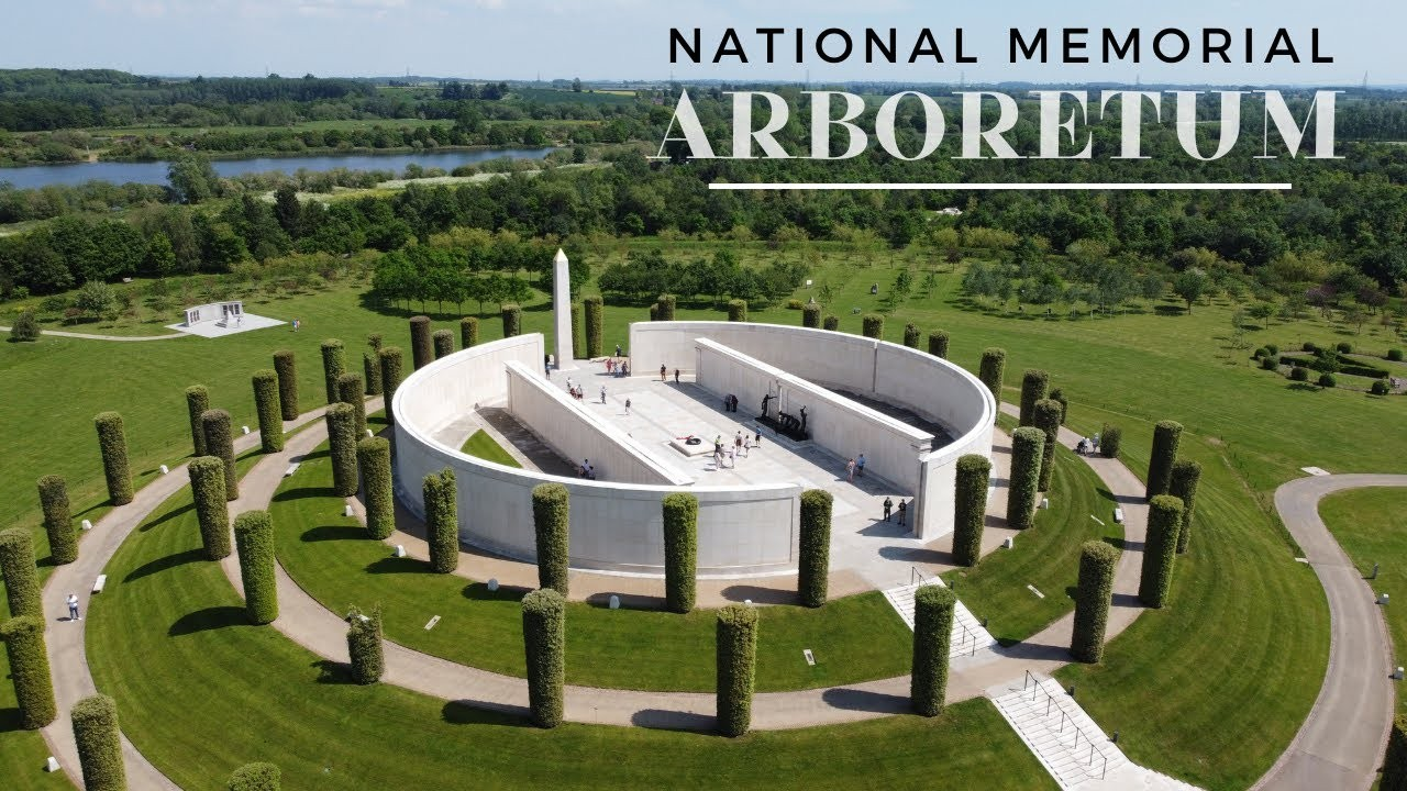 Aerial view of the National Memorial Arboretum featuring a circular white monument with visitors, surrounded by trimmed grass and trees, with a lake and landscape in the background.