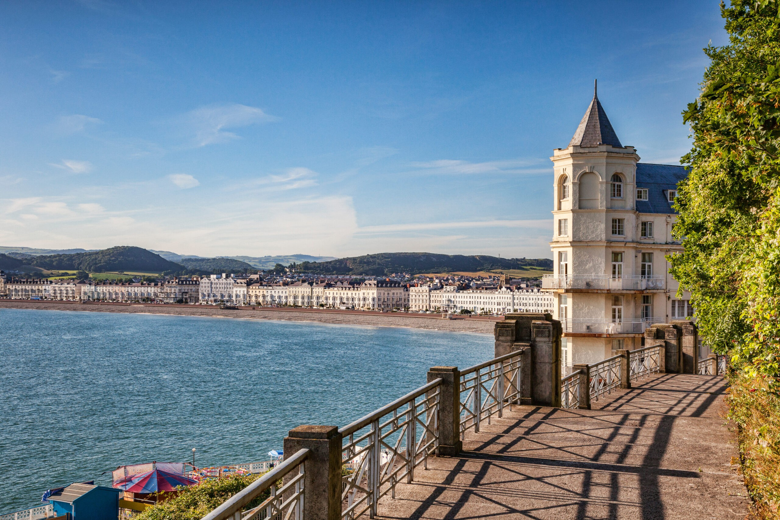 View of a coastal town with white buildings along the shoreline, a large water body in the foreground, a historic tower with a conical roof on the right, and green hills in the background under a clear blue sky.