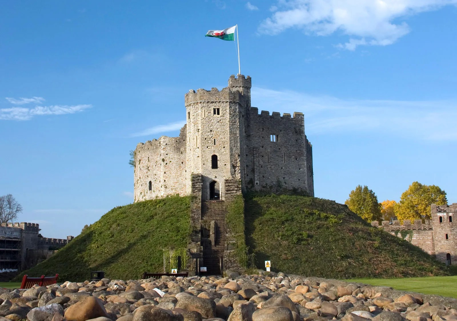 A historic stone castle on a hill with a flag on top, surrounded by a grassy area, rocks, and trees with autumn foliage, under a blue sky.