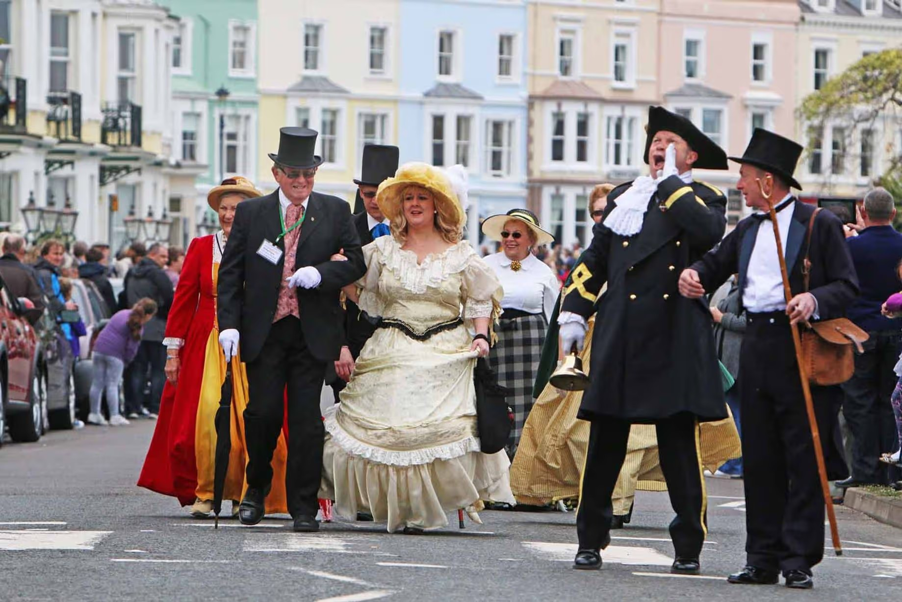 People participating in a parade wearing historical costumes, including a woman in a cream-colored dress and a large hat, a man in a black suit and top hat, and others dressed in 18th or 19th-century attire, with a crowd watching on the sides and colorful buildings in the background.