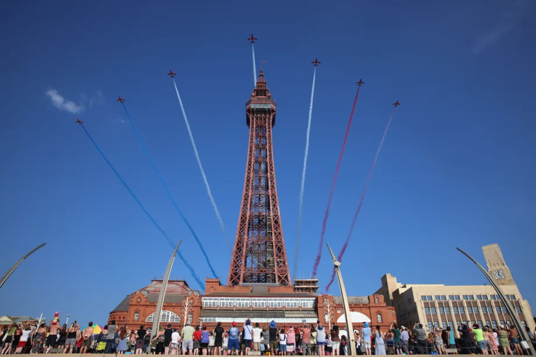 People gathered on an outdoor plaza watching an air show with planes flying around a tall red tower, creating smoke trails in blue, white, and red colors against a clear blue sky.