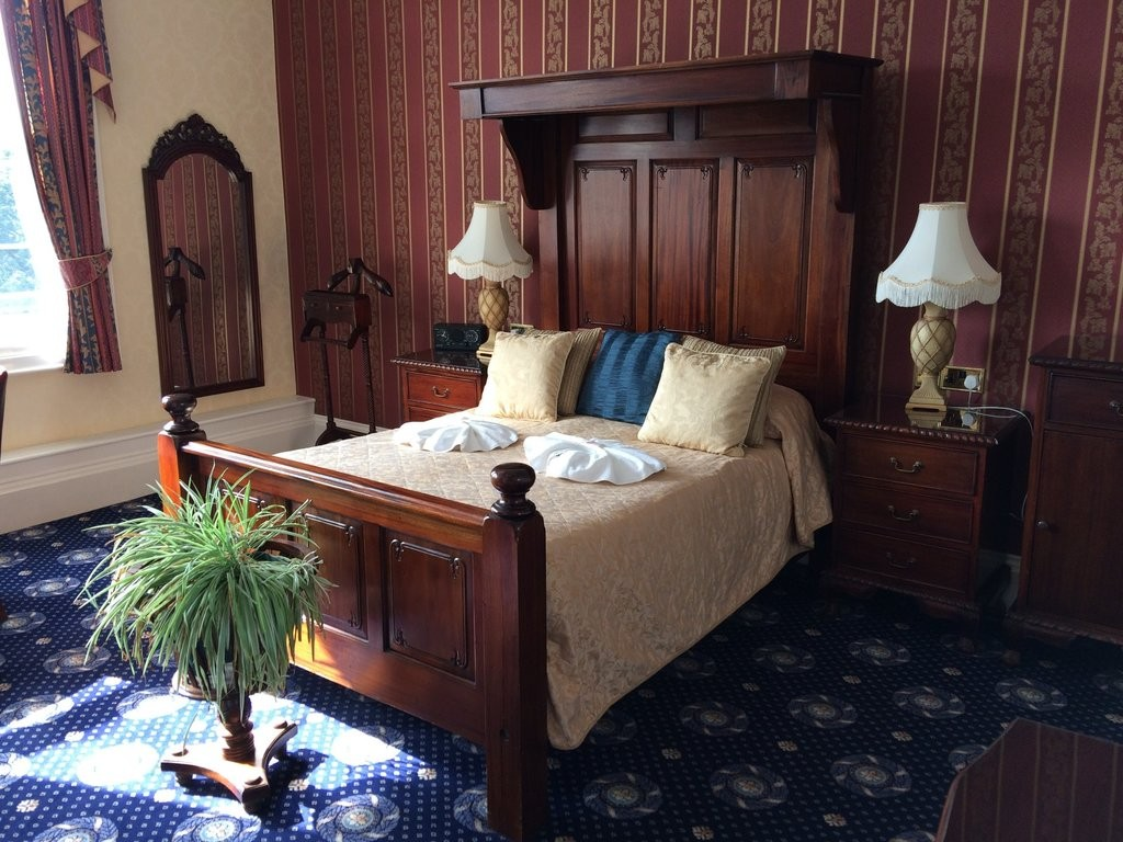 A traditional hotel room with a wooden bed, a tall headboard, and a matching nightstand. The bed has a beige bedspread, two beige pillows, and a blue pillow. There are two lamps on the nightstands with white fringed lampshades. The room has patterned
