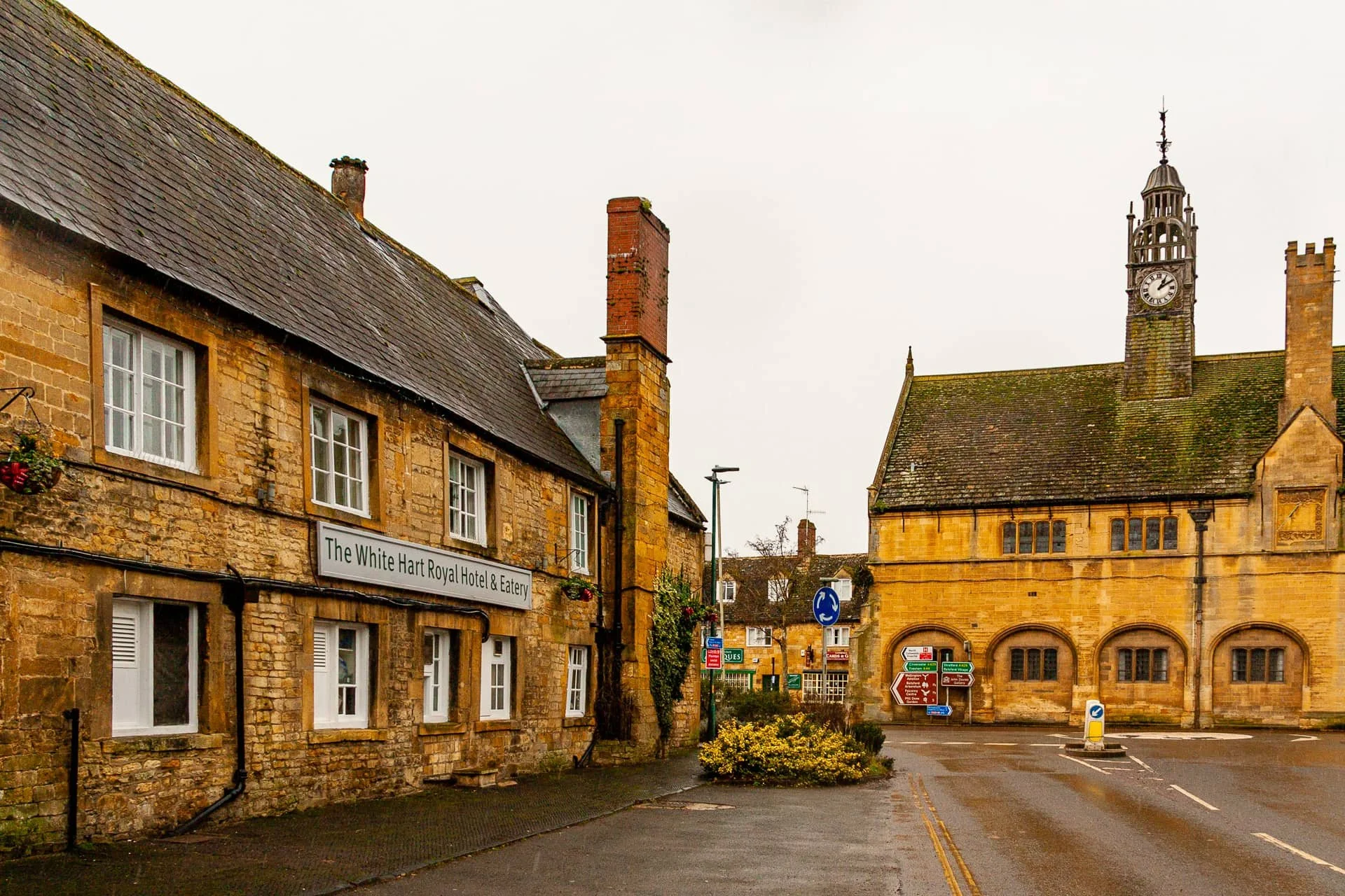 A street scene in a small town with historic stone buildings, featuring a hotel and a church with a clock tower, overcast skies, wet pavement, and street signs.