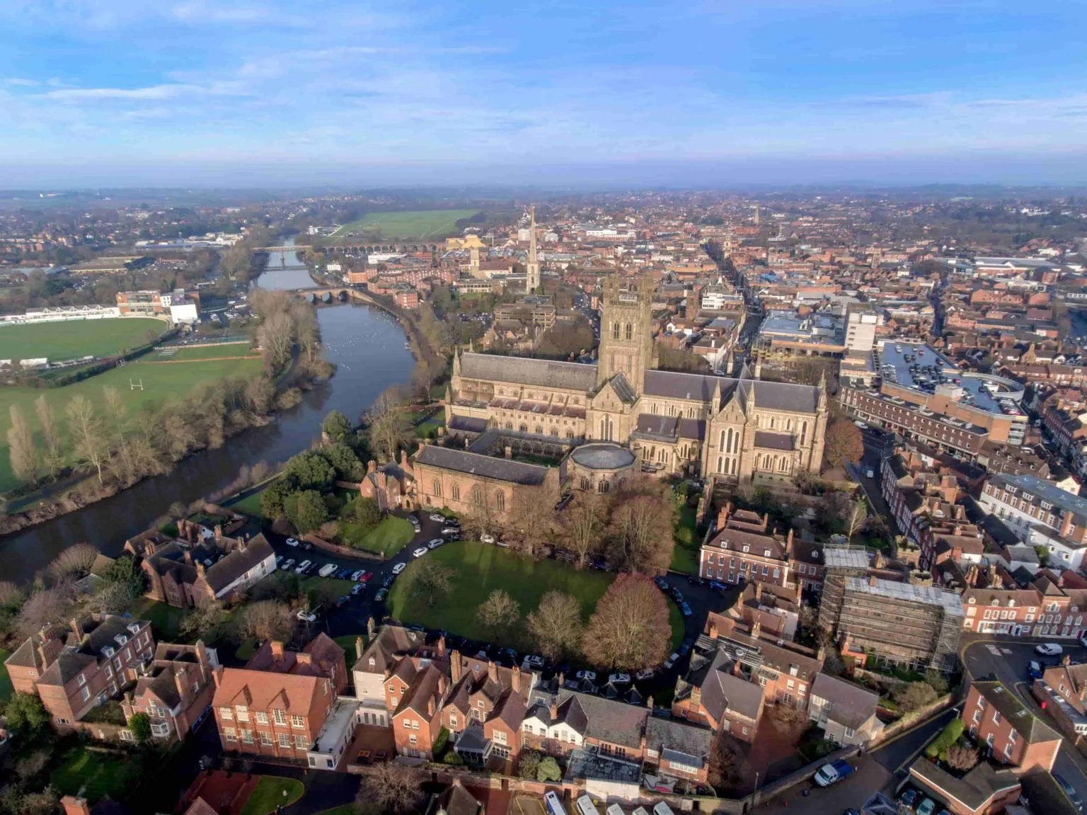 Aerial view of a historic church with a tall tower, surrounded by a river, parks, and city buildings under a blue sky in a town.